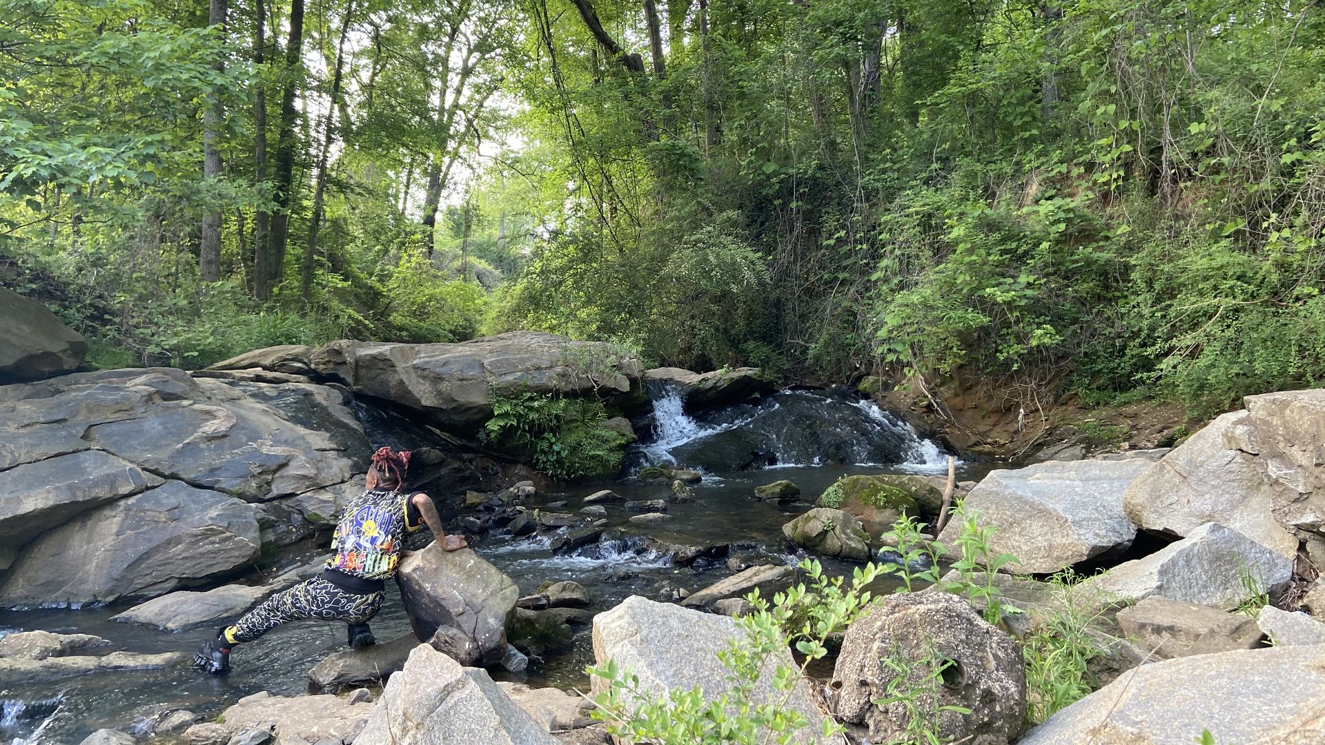 A person stretches their leg to reach a rock in a rushing creek surrounded by green trees