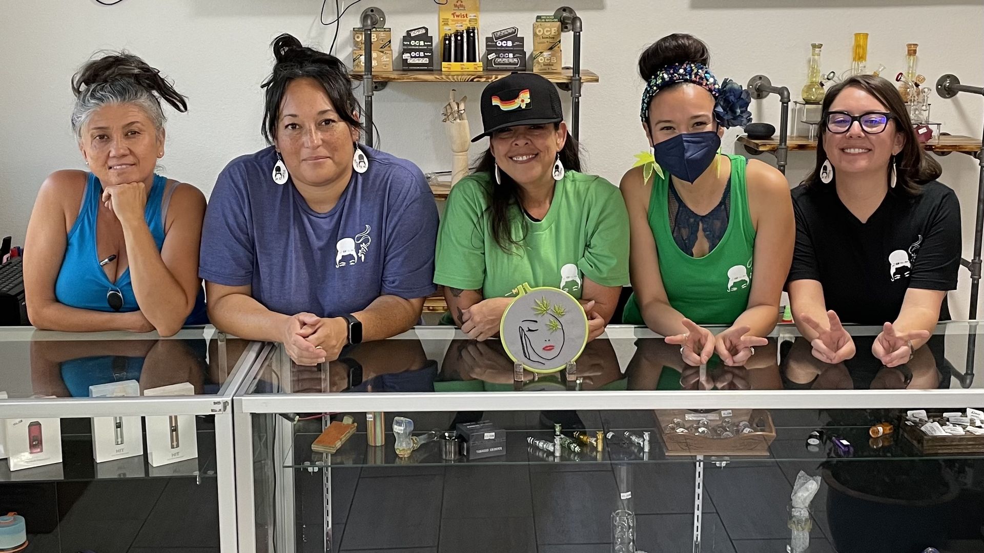Five women pose while leaning on a glass case full of marijuana accessories, like glass pipes