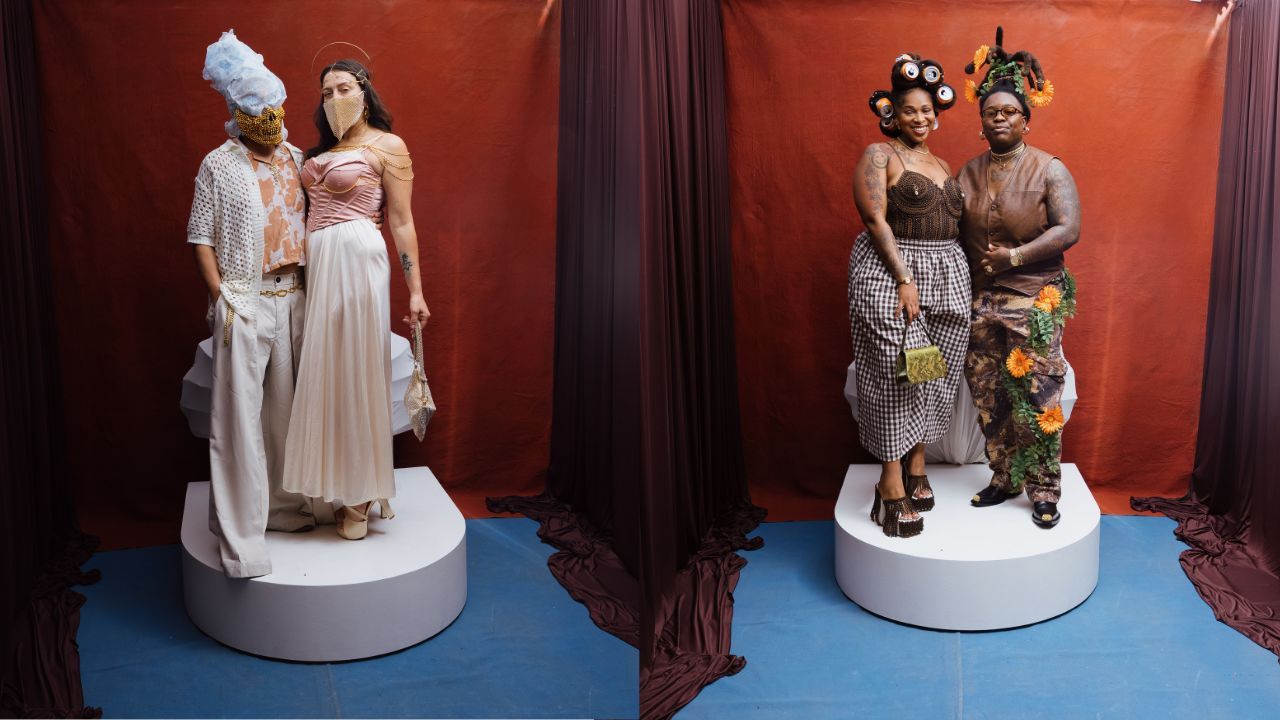 Two fashion displays on white pedestals before a red backdrop. Left: a person with a blue headpiece and gold mask beside a pale pink gown. Right: two people in bold patterns pose.