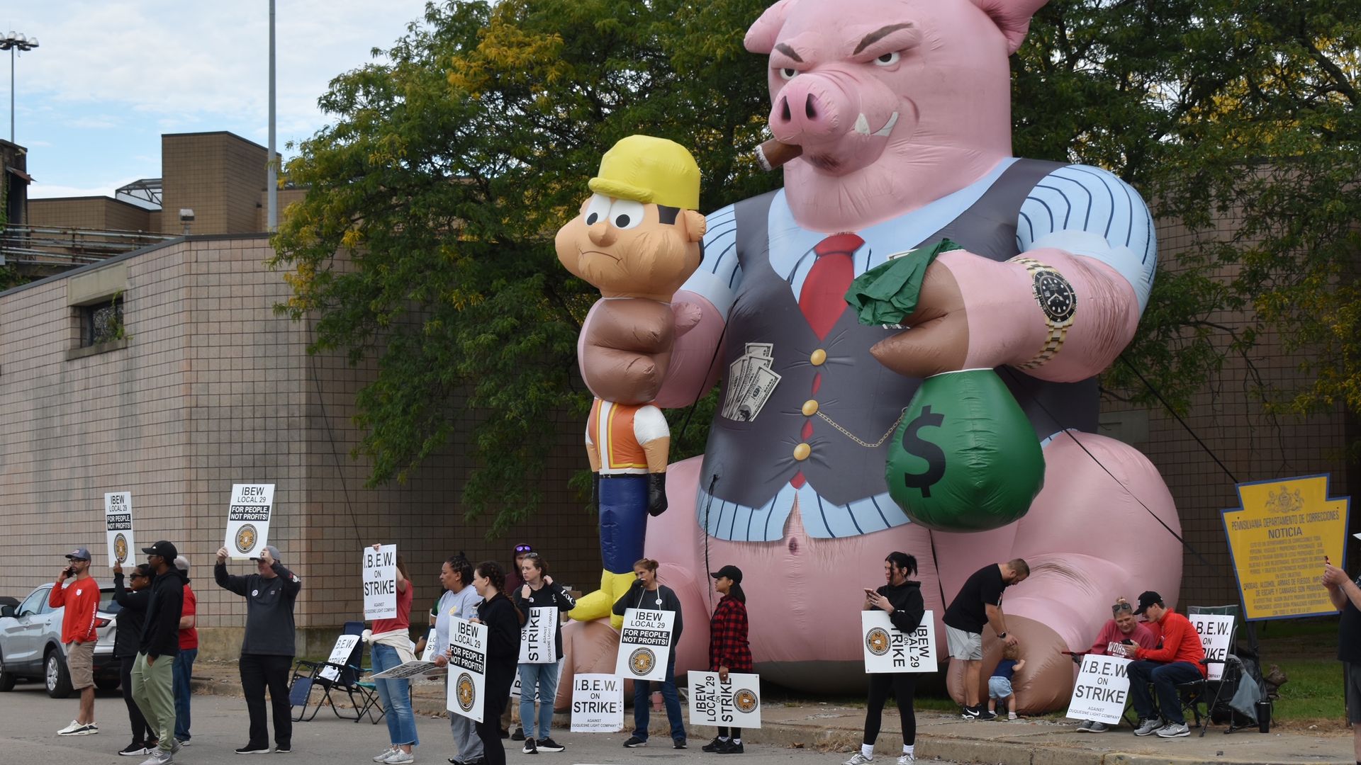Massive pink inflatable pig in a vest stands beside a smaller inflatable man in a yellow hard hat; crowd of protesters holds IBEW strike signs before a brick building.