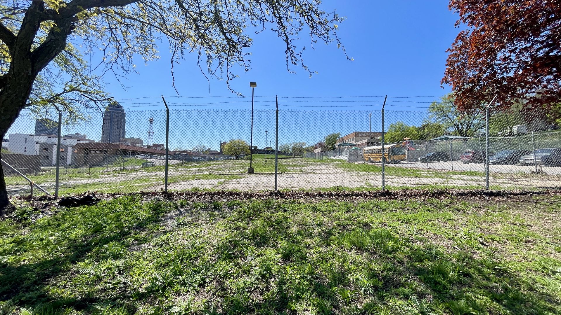 Sunny city lot behind a chain-link fence topped with barbed wire; a yellow school bus sits among low buildings, with a tall skyscraper in the distance, and green grass and trees framing the scene.