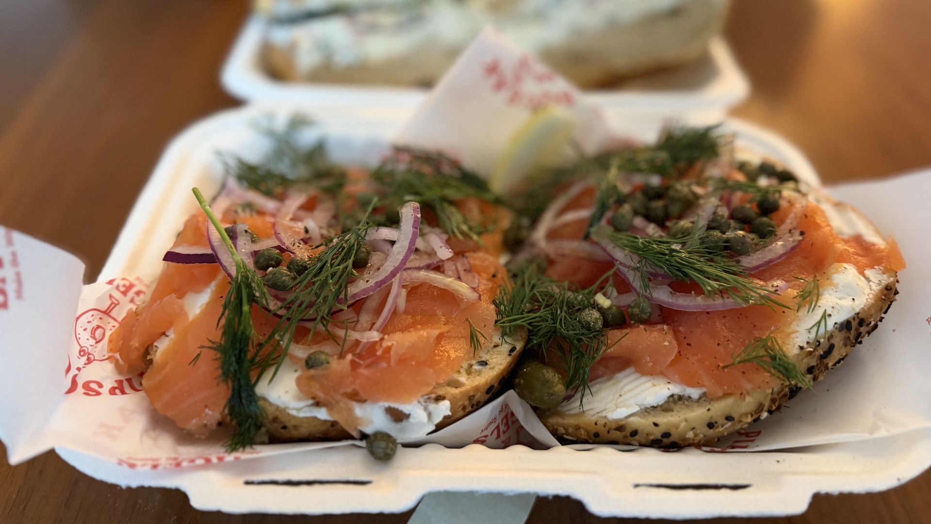 Two open-faced sesame bagel halves topped with cream cheese, smoked salmon, red onion, dill, and capers, with a lemon wedge; blurred bread in the background.