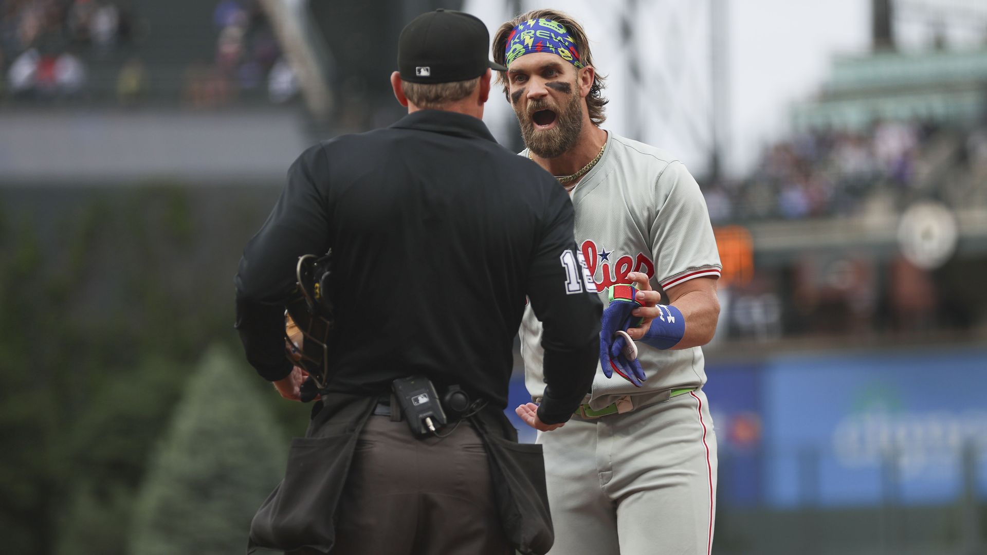 Baseball player wearing a gray Phillies uniform and colorful headband argues with umpire dressed in black during a game, displaying an intense expression and eye black under his eyes.