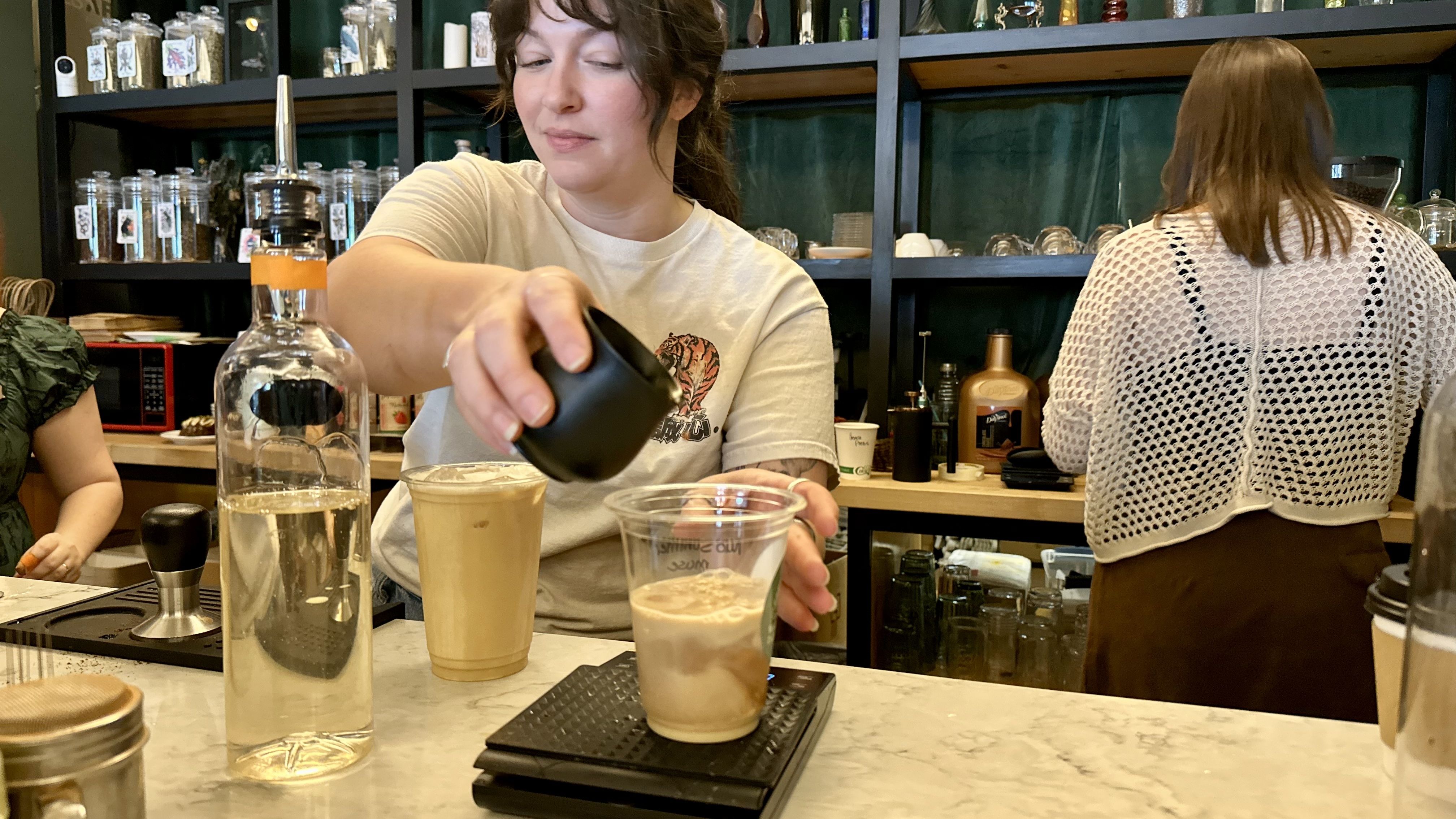 Barista with brown hair pours from a black pitcher into a cup of coffee at a busy cafe counter, with jars and bottles on shelves in the background and another person facing away.
