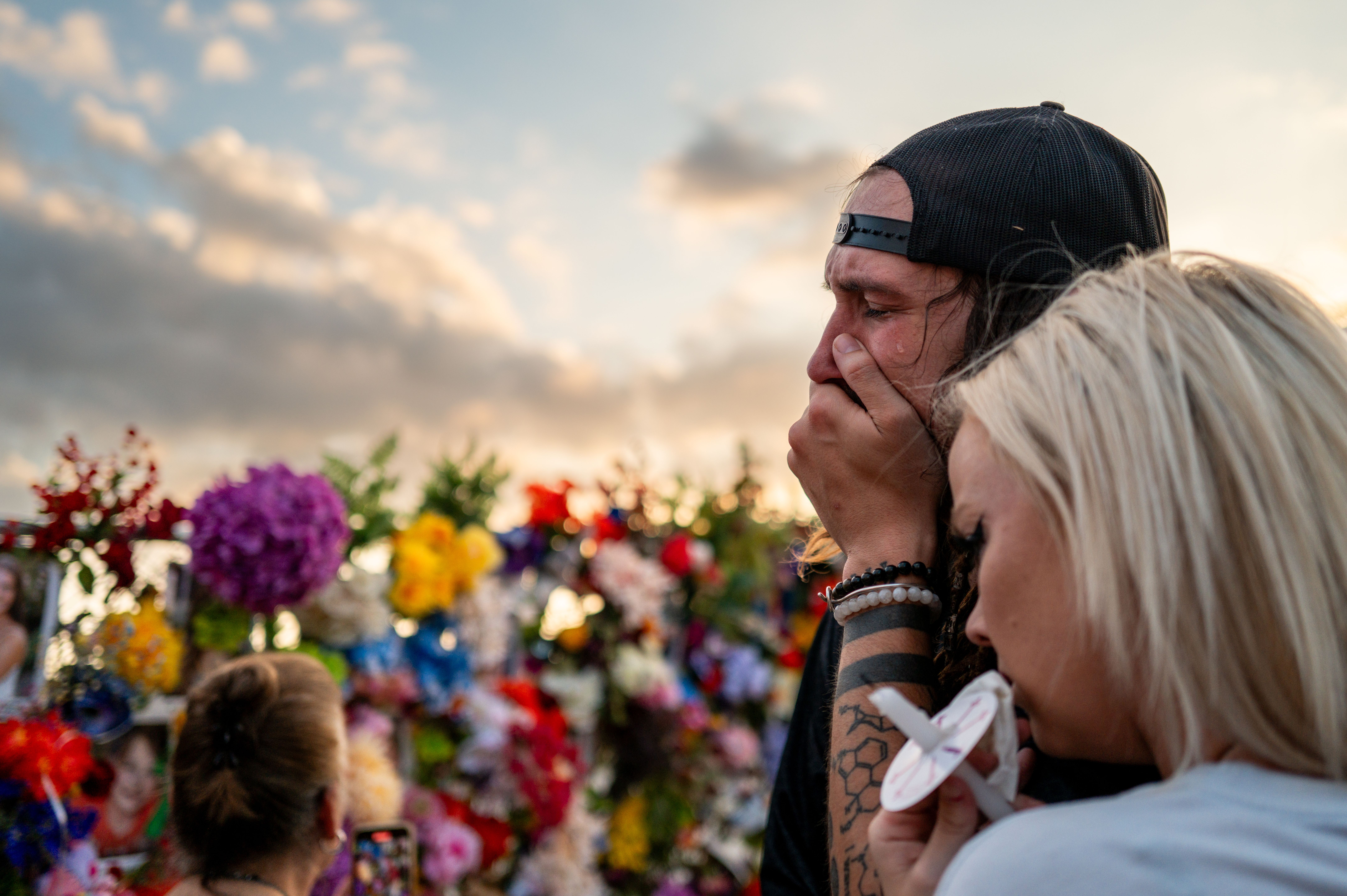 Community members grieve during a candlelight vigil for those who died in the July Fourth, 2025 flooding in the Texas Hill Country. A man in a black baseball cap is seen from the side crying and holding a hand to his mouth, while a woman comforts him. Flowers are blurred in the background.