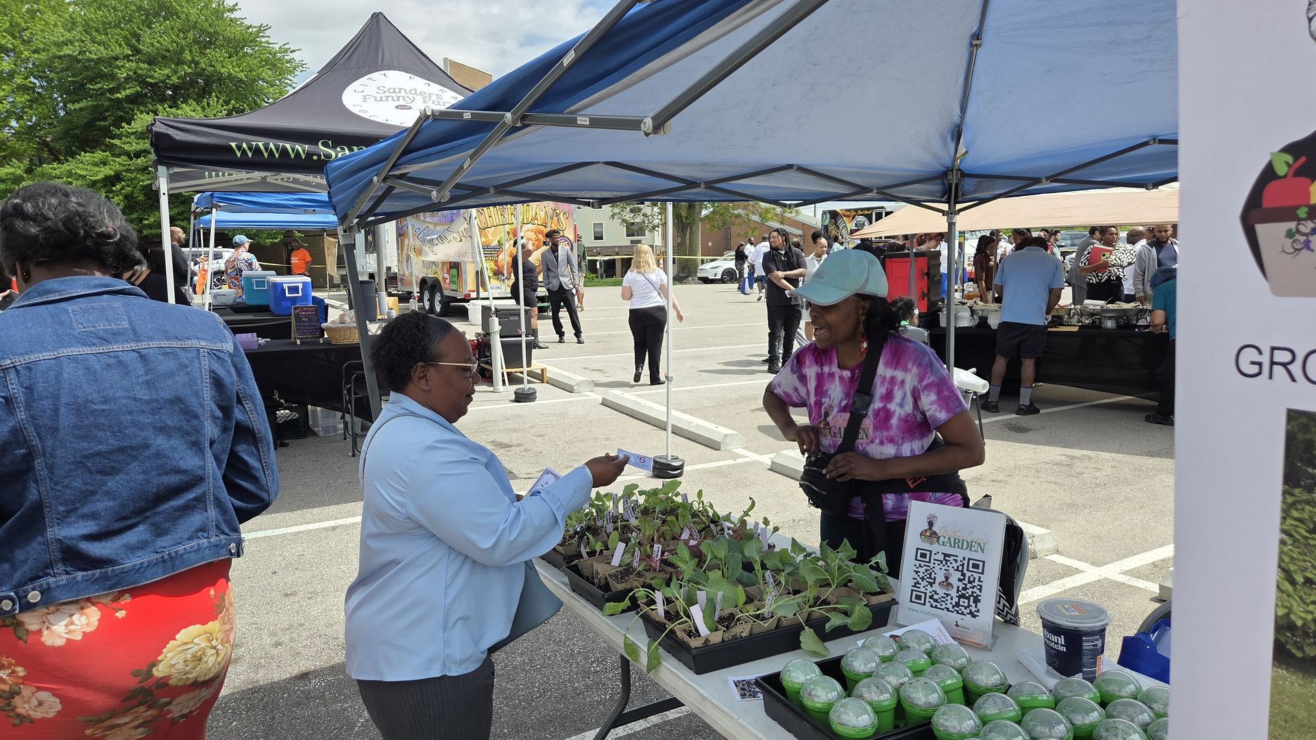 Outdoor market under blue canopies; a woman in a blue blouse chats with a vendor at a seedling table, while another woman in a purple tie-dye shirt smiles. Food trucks and booths in the background.