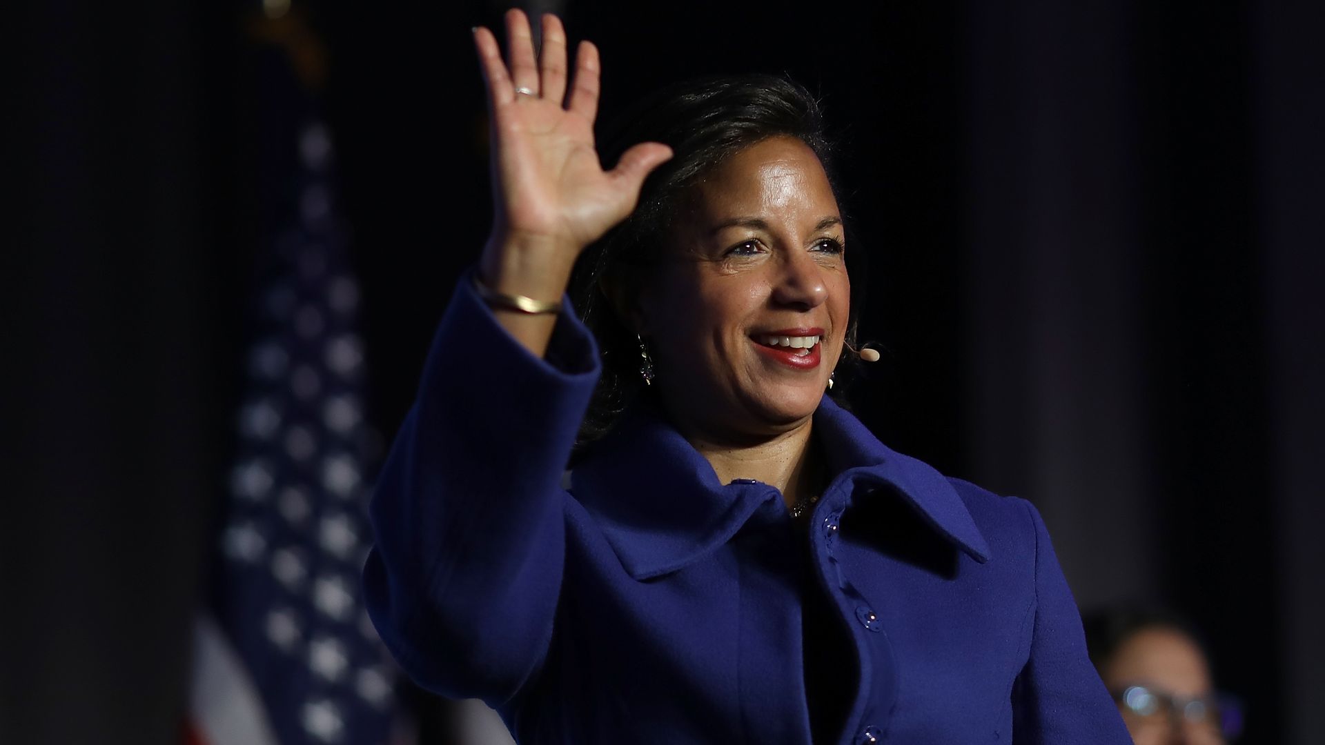Former National Security adviser Susan Rice smiles and waves to a conference audience with an American flag in the background.