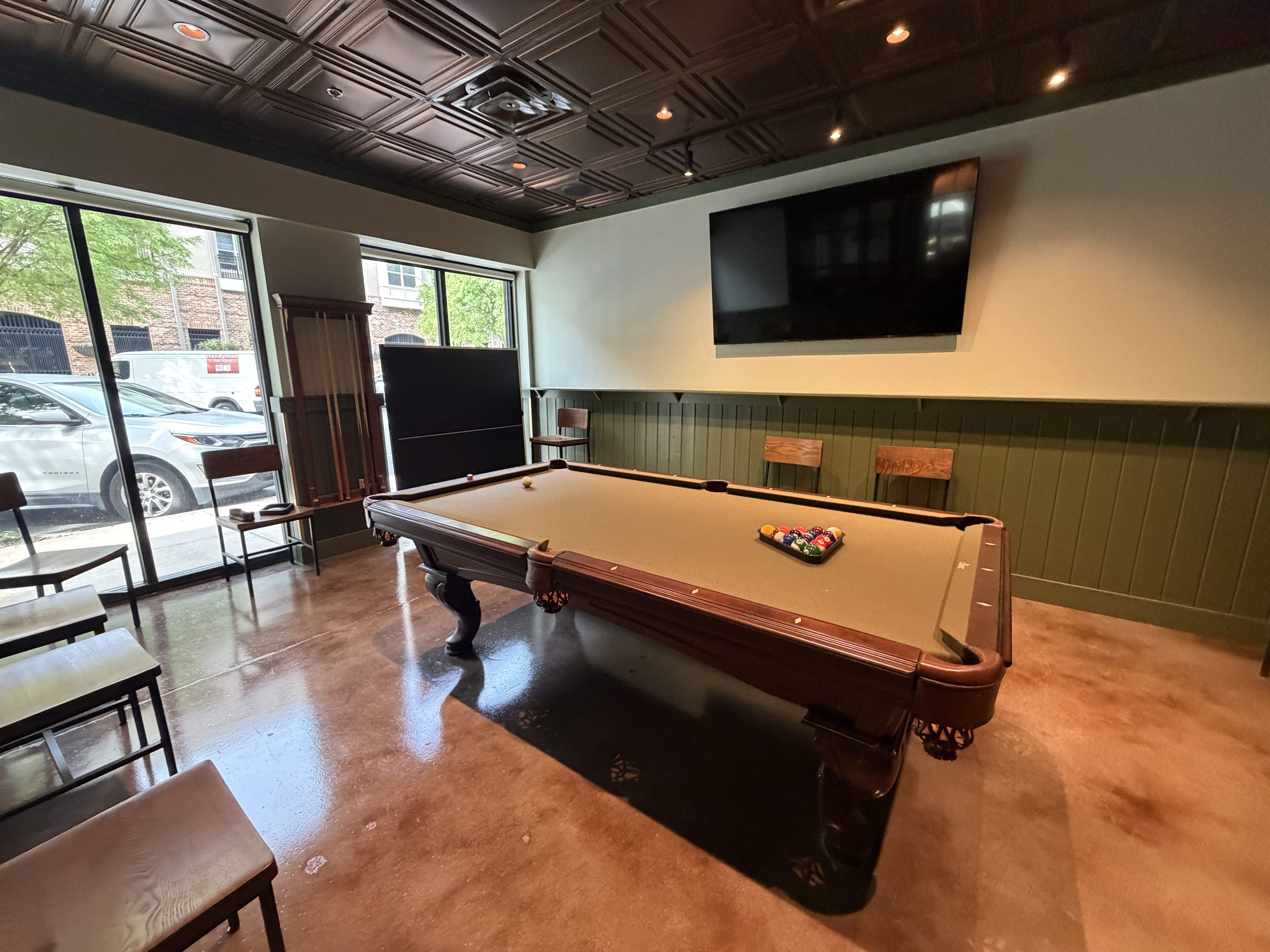 Indoor billiards room with a dark wood pool table in the center, colorful balls near the far end, chairs along the walls, a large wall TV, green wainscoting, and windows showing parked cars outside.