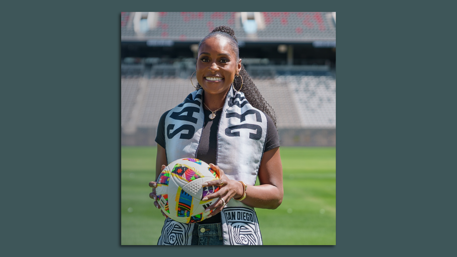 Actress Issa Rae wears a San Diego FC scarf and stands on the field holding a soccer ball. 