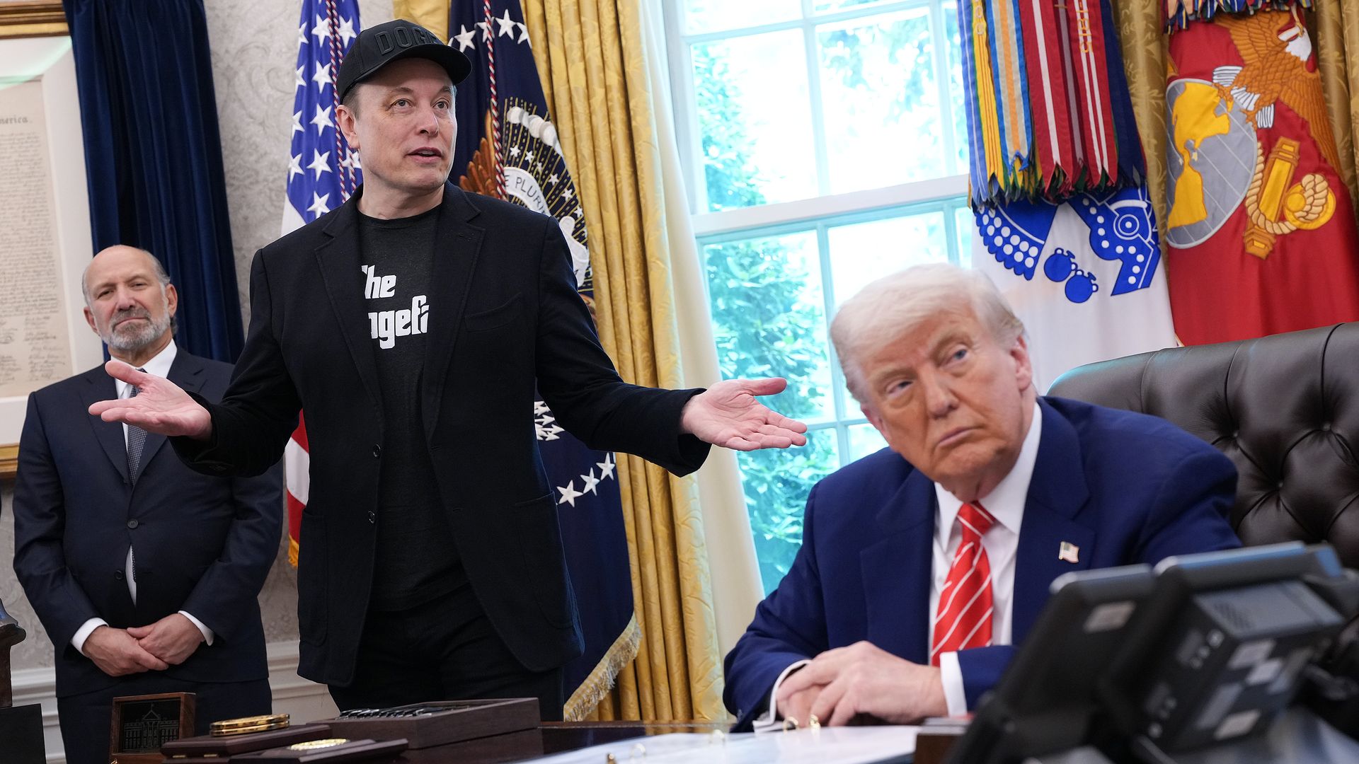 Elon Musk wearing a black blazer and hat, while flanked by Donald Trump and Howard Lutnick in the Oval Office.