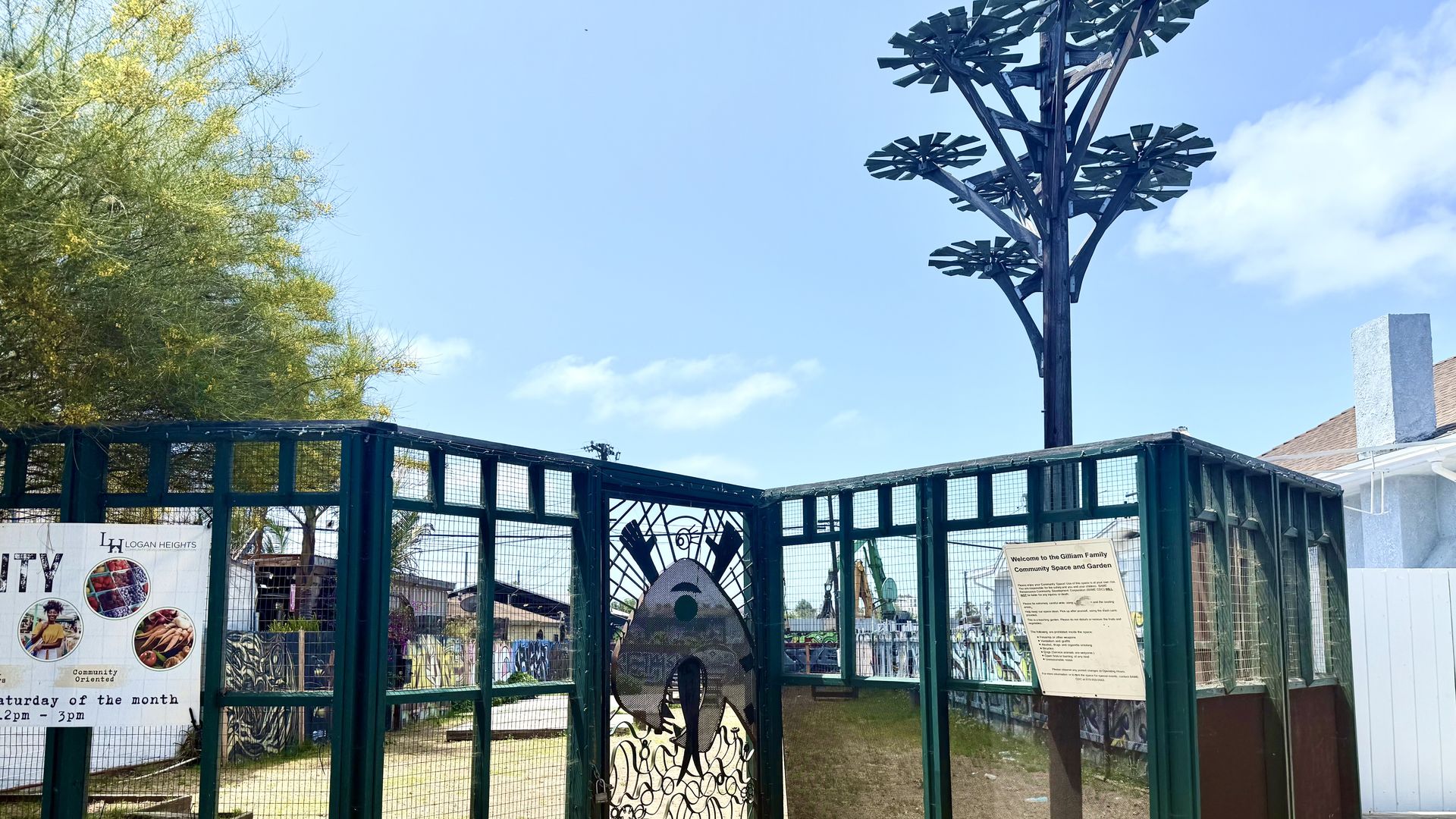 A fence in front of a dirt lot with interior fences filled with grafiti and a wooden fake tree used for shade