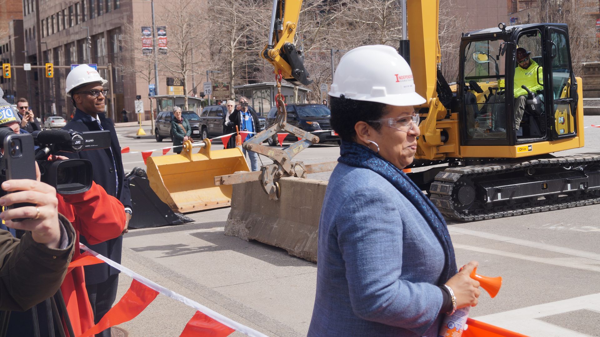 City street construction scene with a large yellow excavator, operator in bright vest, white hard hats worn by workers, onlookers filming, orange safety tape, tall buildings in background.