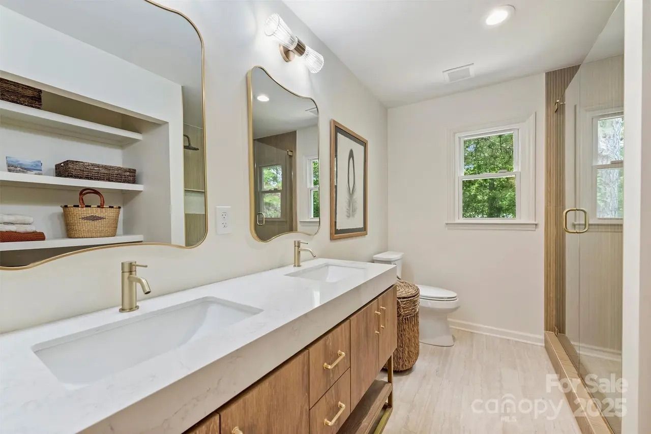 Modern bathroom with double sinks, wooden vanity, gold faucets, two asymmetrical mirrors, woven basket, toilet, glass shower, and window showing green trees outside.