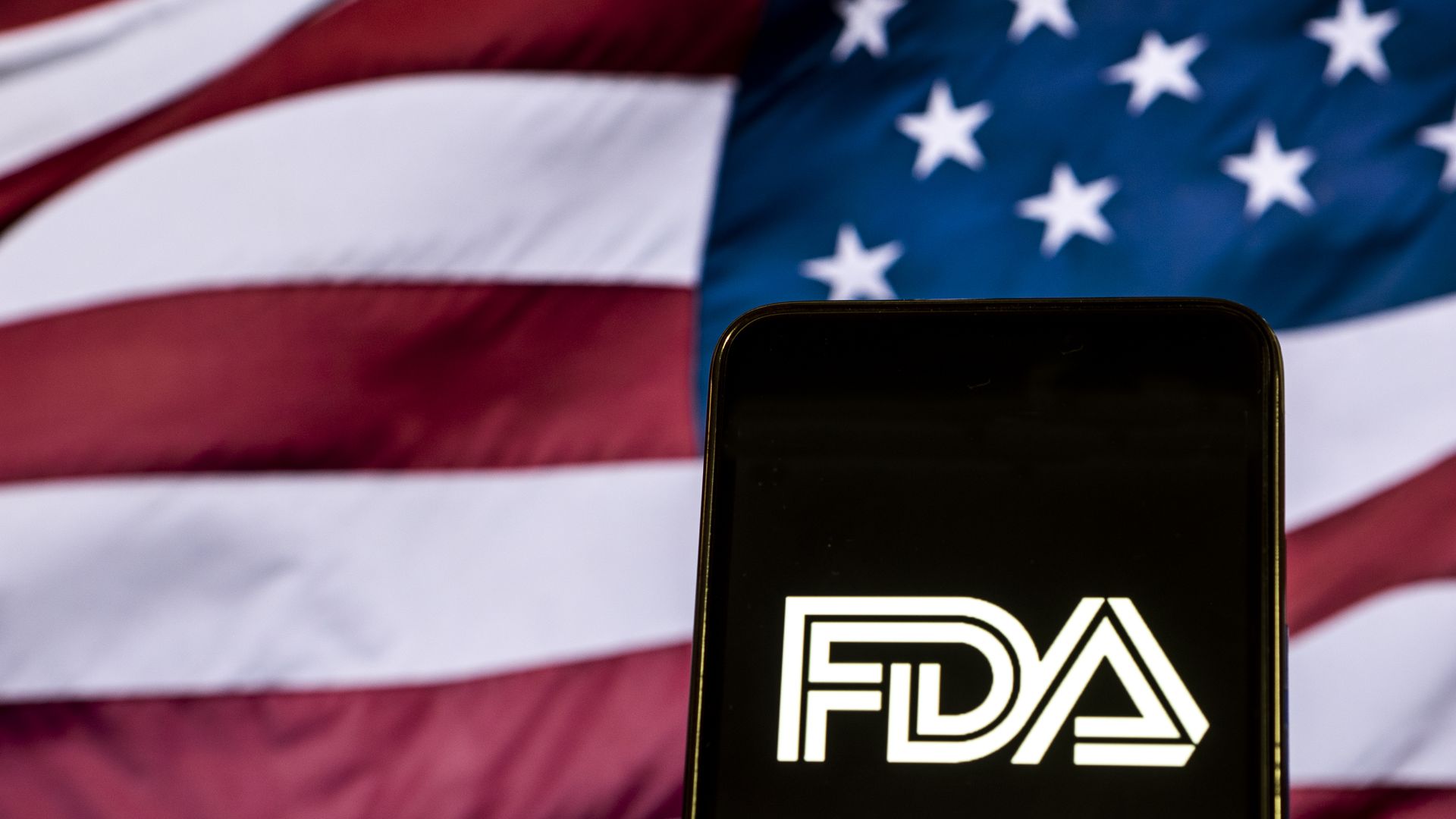 A phone holds up the FDA logo, an American flag waves in the background.
