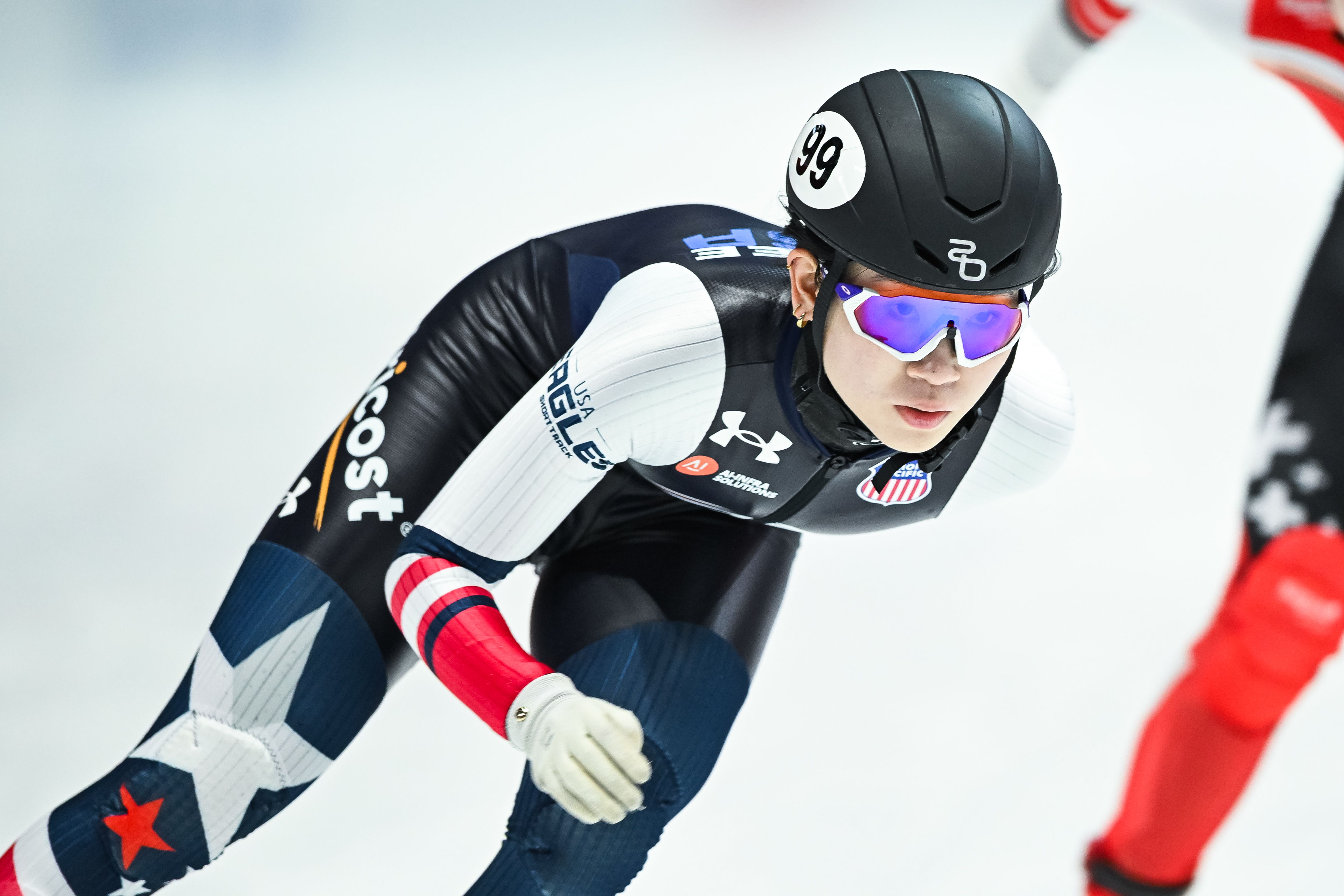 Female speed skater in black helmet with number 99, reflective purple sunglasses, and USA-themed racing suit in black, white, blue, and red, focused on the ice during a race.