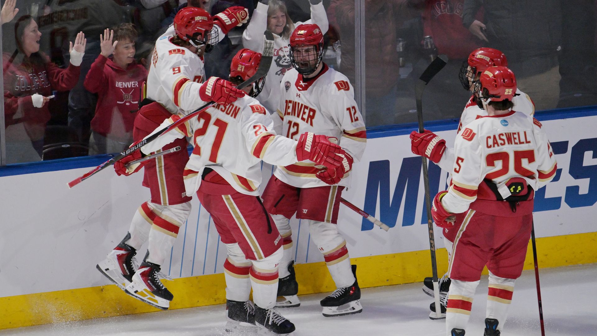 Denver hockey players in white and red celebrate at the boards after a goal, high-fiving and skating, as cheering fans in red raise hands behind the glass.