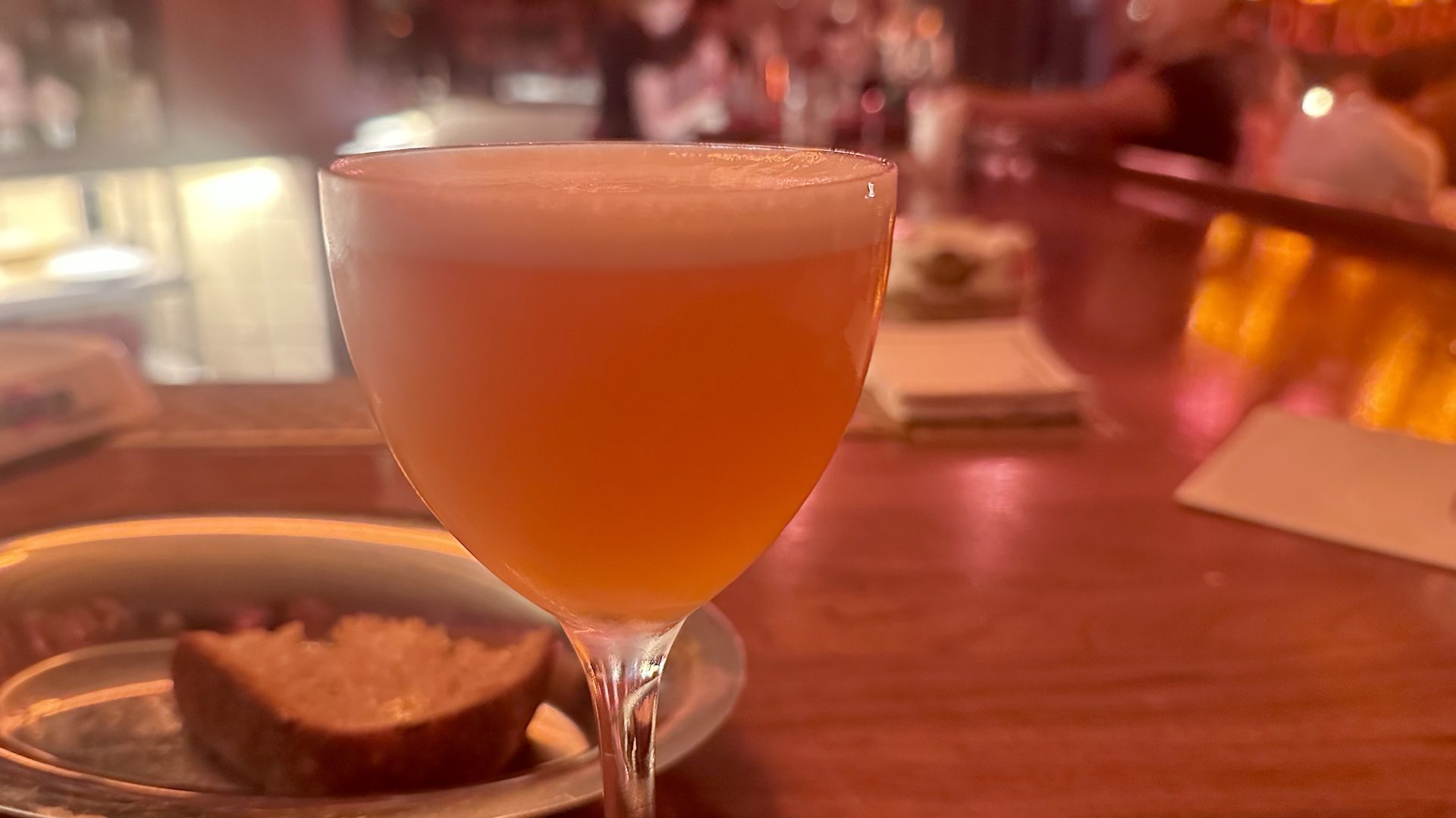 Close-up of a frothy pink cocktail in a stemmed glass on a wooden bar counter with a plate holding a piece of bread in a warmly lit bar setting.