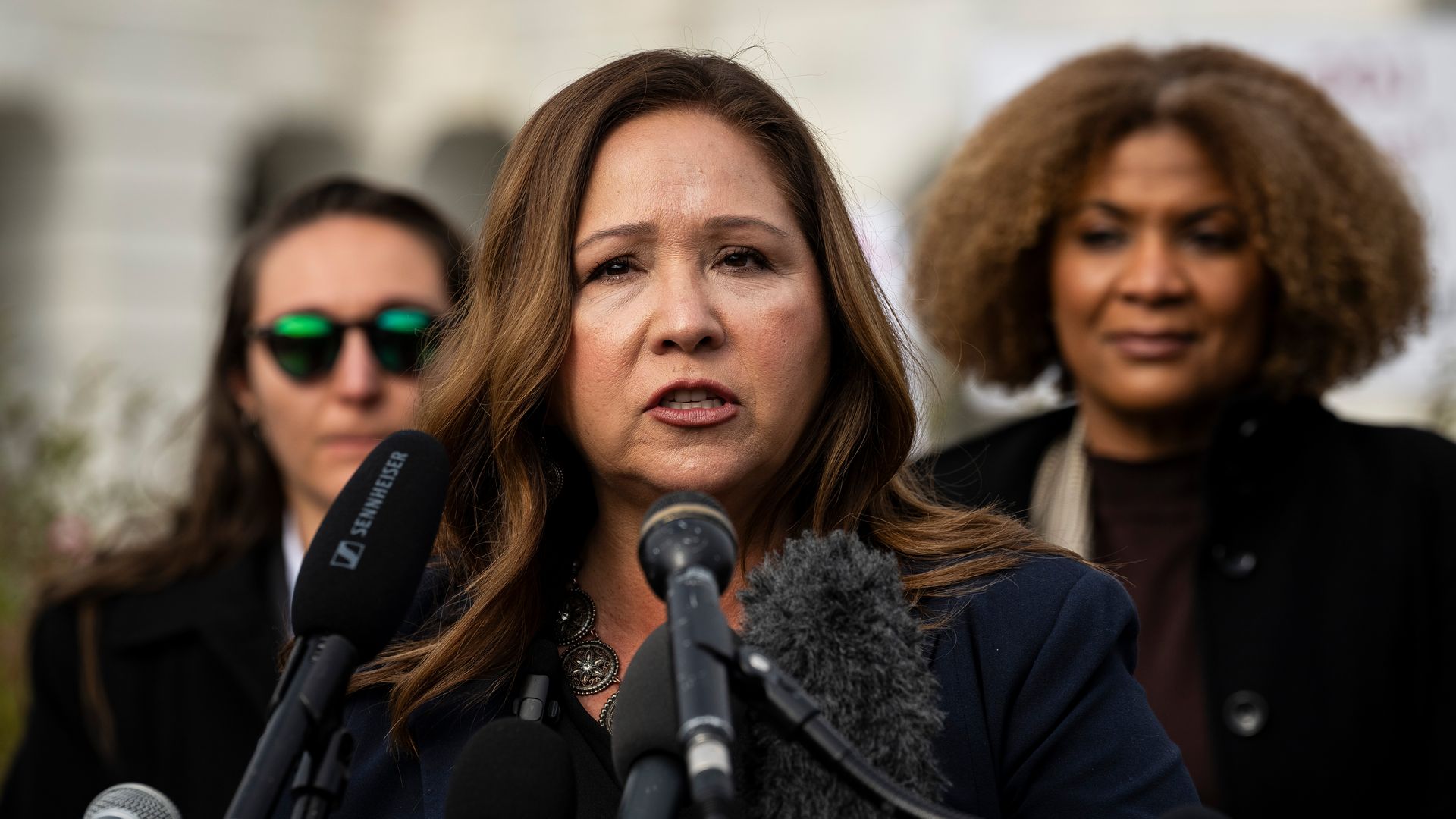 Rep. Adelita Grijalva, wearing a blue suit and a black blouse, speaks into microphones in front of a row of people and the U.S. Capitol.