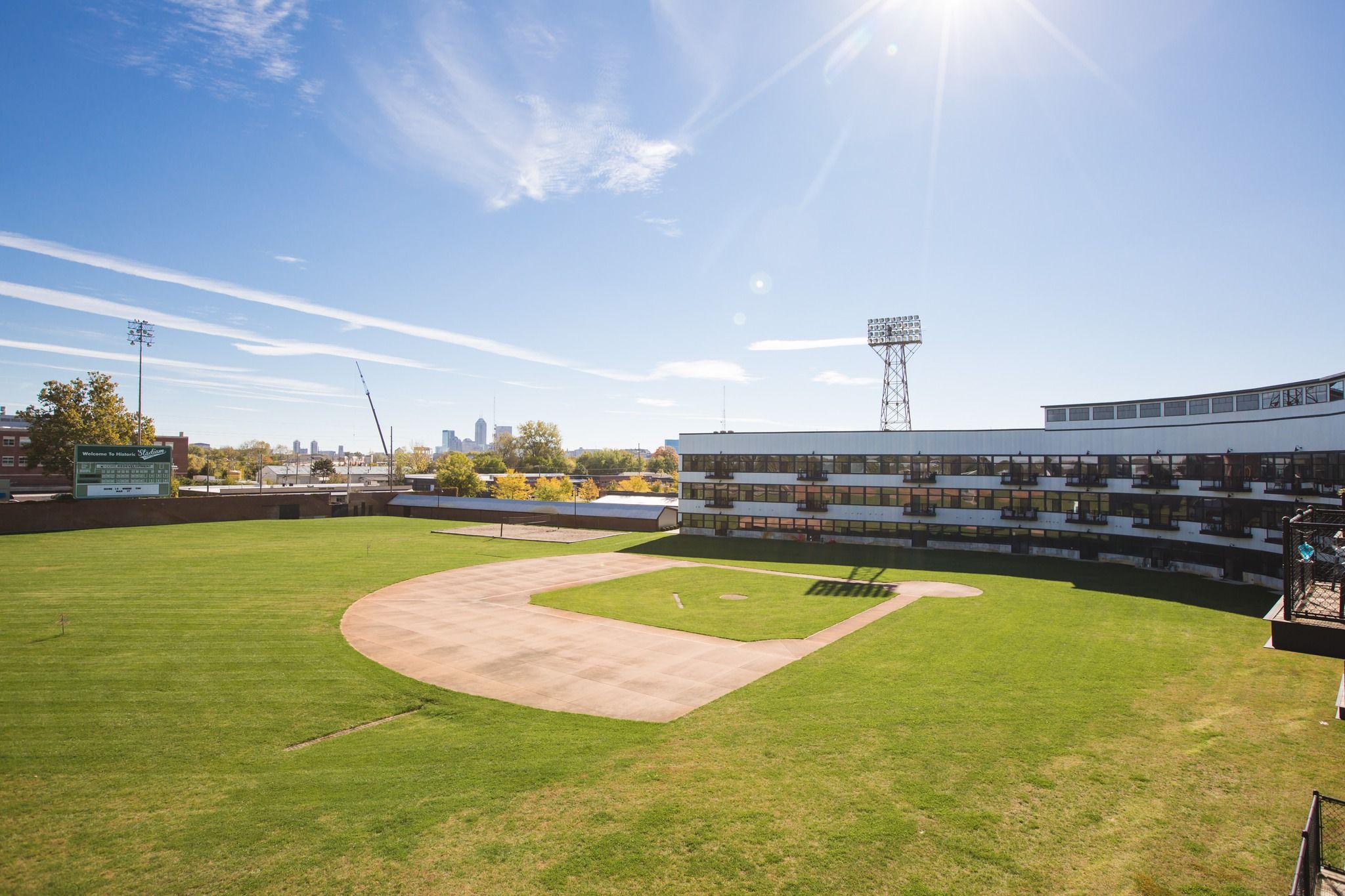 Empty historic baseball field with green grass, a dirt infield, a scoreboard, stadium lights, surrounded by a building with glass windows under a clear blue sky with the sun shining.