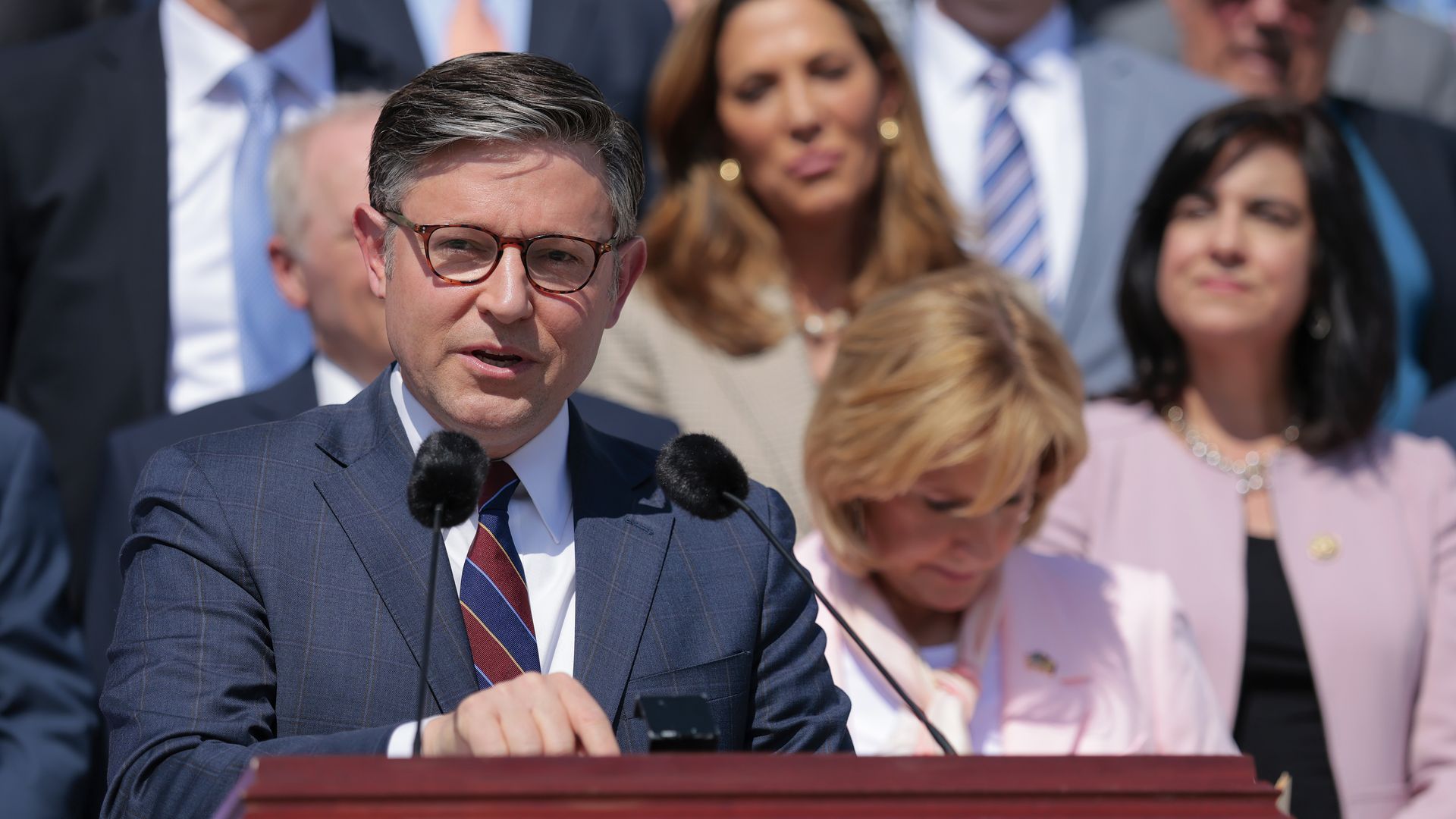 A man in a blue check suit and red striped tie speaks at a wooden podium with two microphones, surrounded by smiling people in business attire.