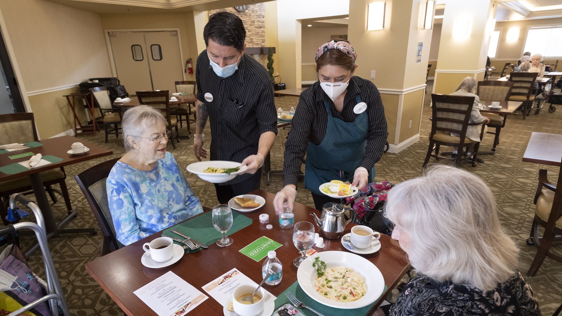 Two workers serve food to two elderly women at a senior living center.