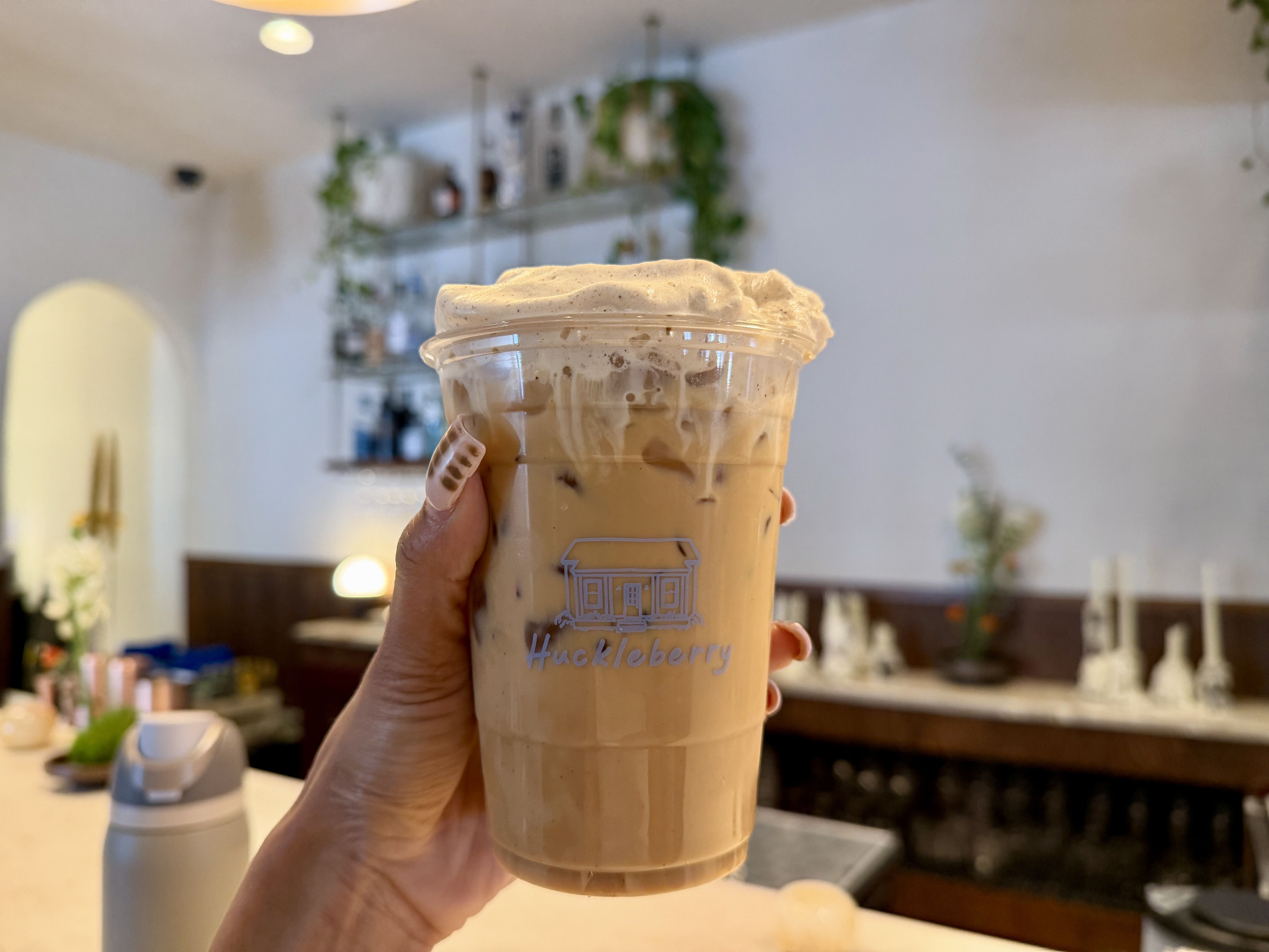 Close-up of a hand holding a tall iced coffee with whipped cream in a clear cup; cafe interior with shelves and green plants blurred in the background. Huckleberry Noda.