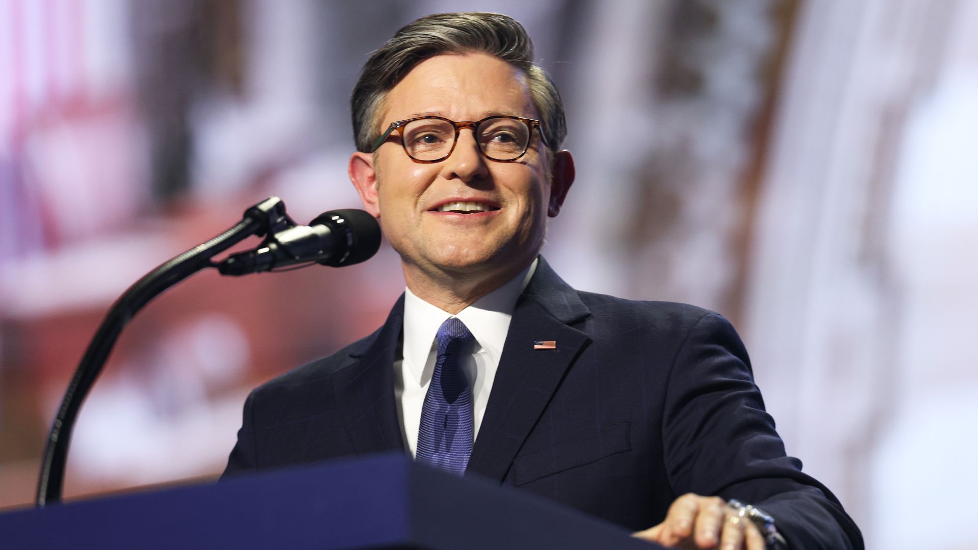 A man in a dark suit and blue tie stands at a podium with a microphone, wearing tortoise-shell glasses and a small American flag pin; smiling, with a blurred crowd in the background.