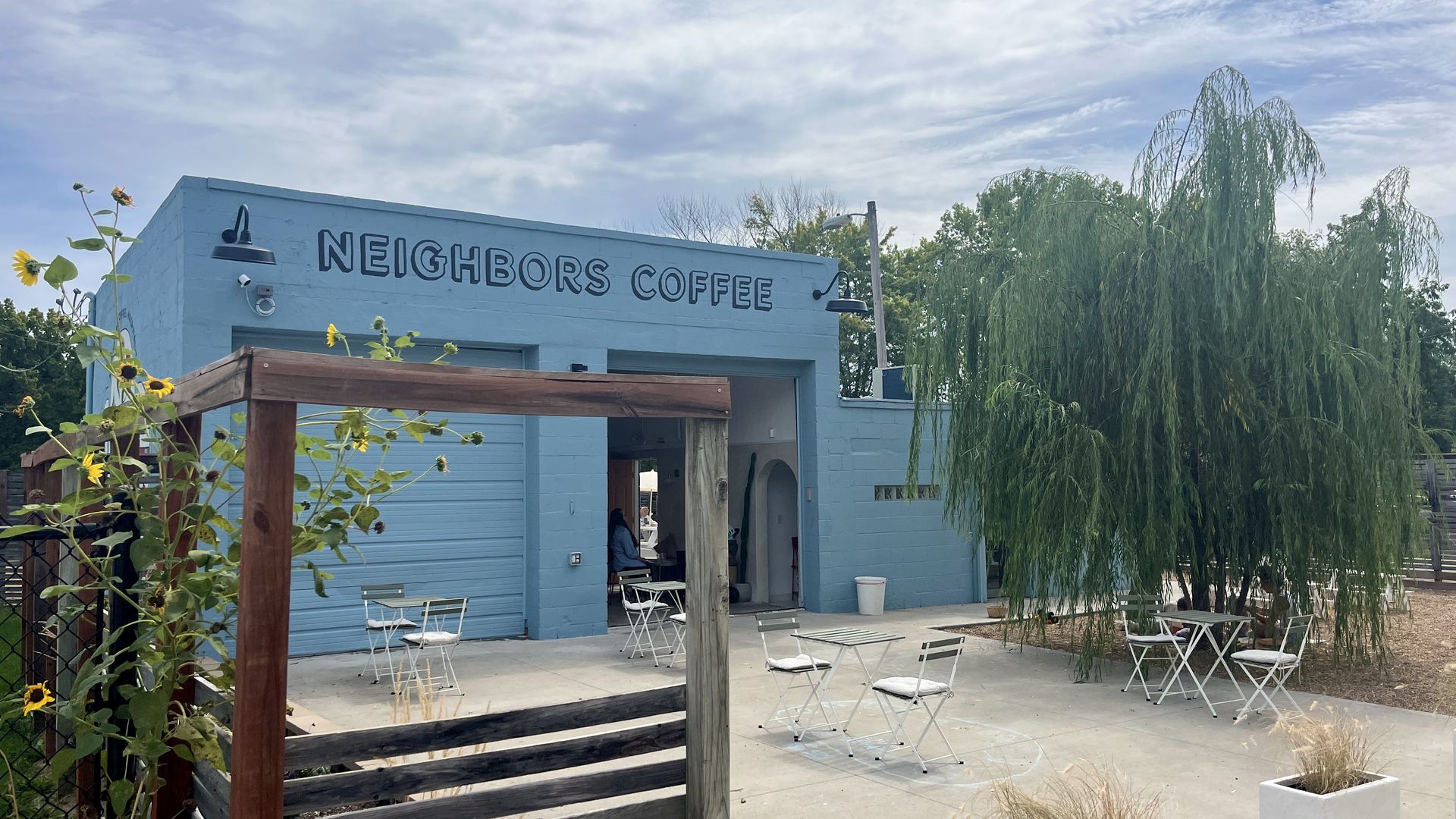 Blue building with "Neighbors Coffee" sign, outdoor seating with white chairs and tables, sunflowers on wooden trellis, and a large green tree under a partly cloudy sky.