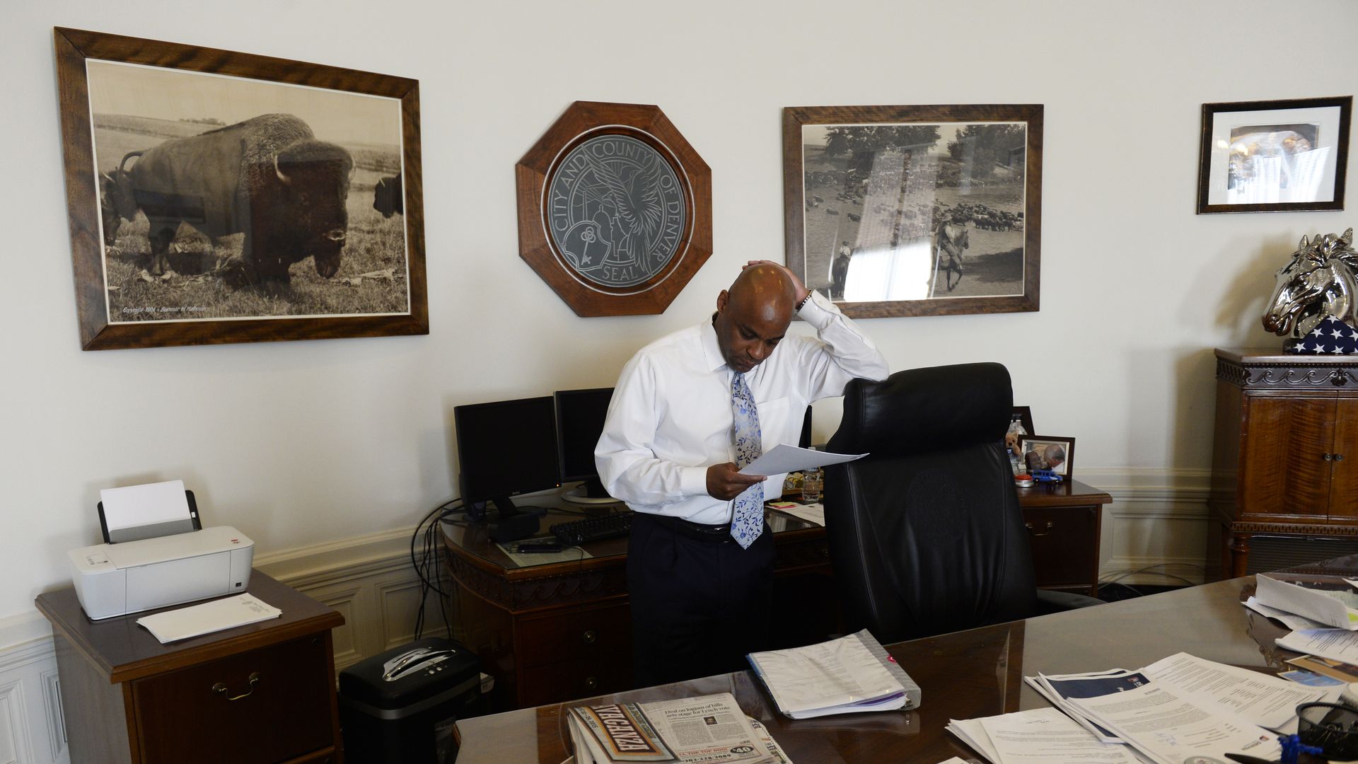 A photo of Denver Mayor Michael Hancock standing at his desk and looking down at some documents with his hand on his head.