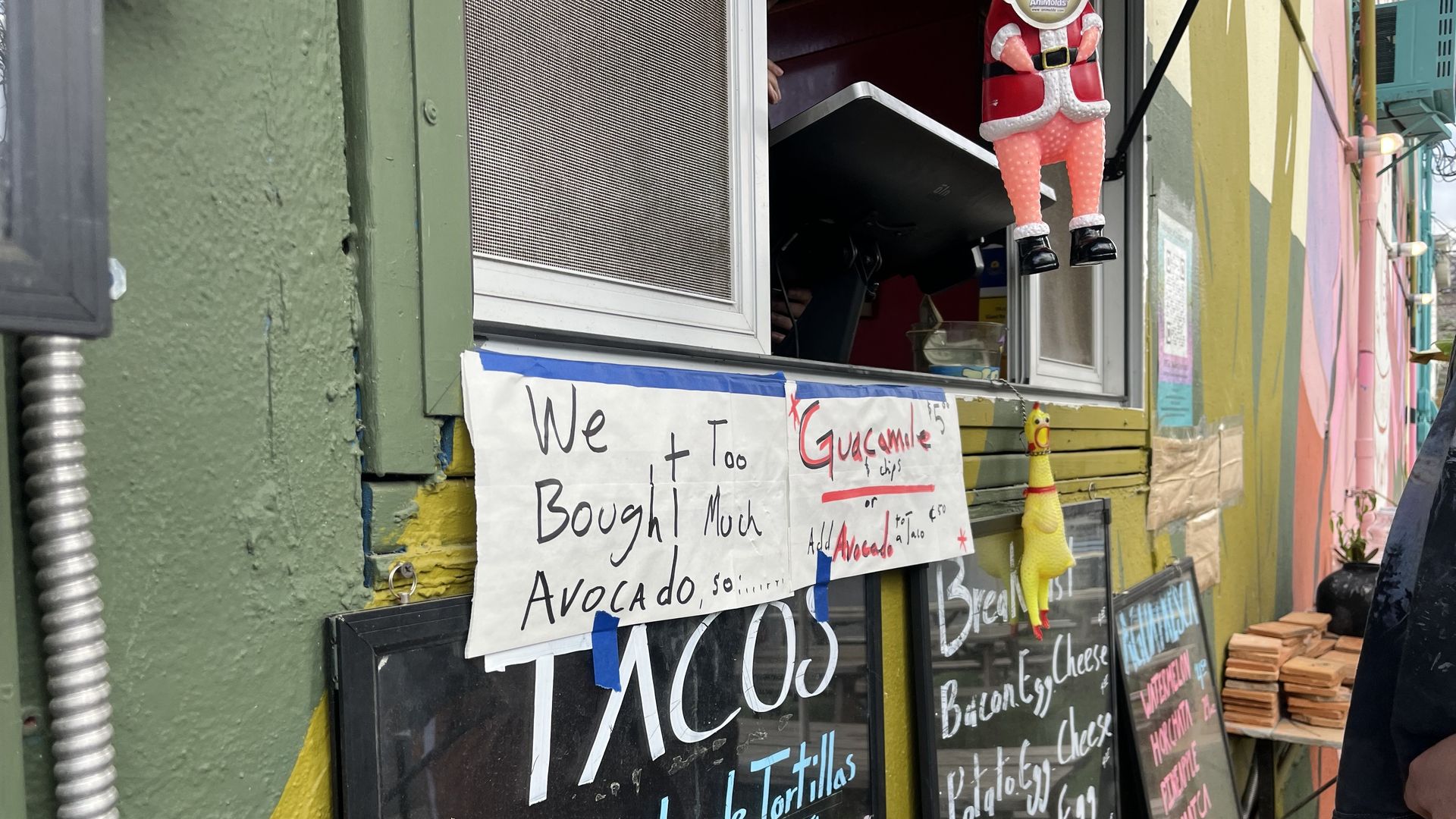 Colorful storefront with green and yellow walls, outside chalkboard menus, and a handwritten sign reading "We Bought Too Much Avocado". Hanging decorations include a Santa figure and a yellow giraffe.