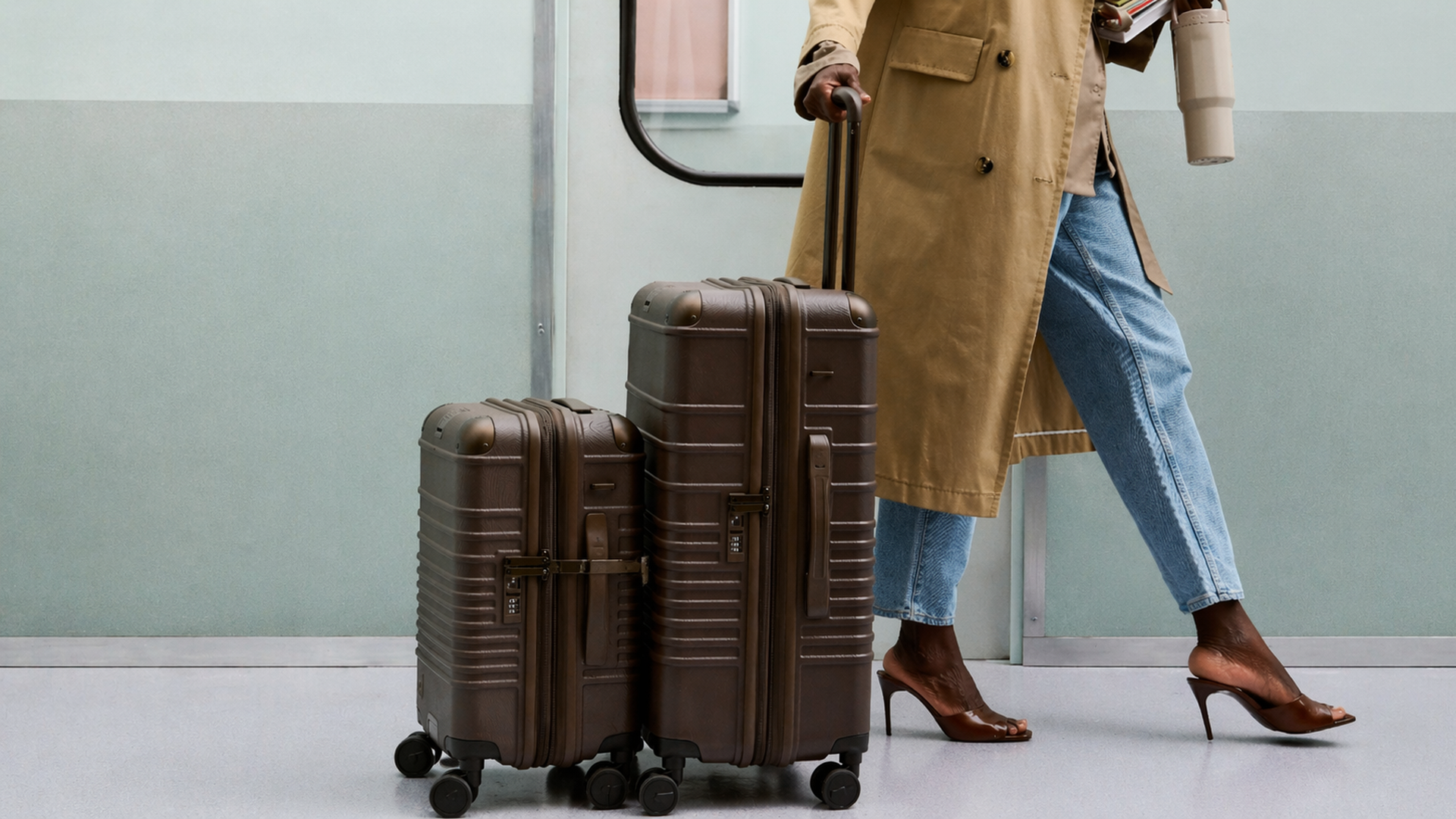 A person in a beige trench coat walks with two brown rolling suitcases; wearing blue jeans and brown heeled sandals, they hold a coffee cup and documents.