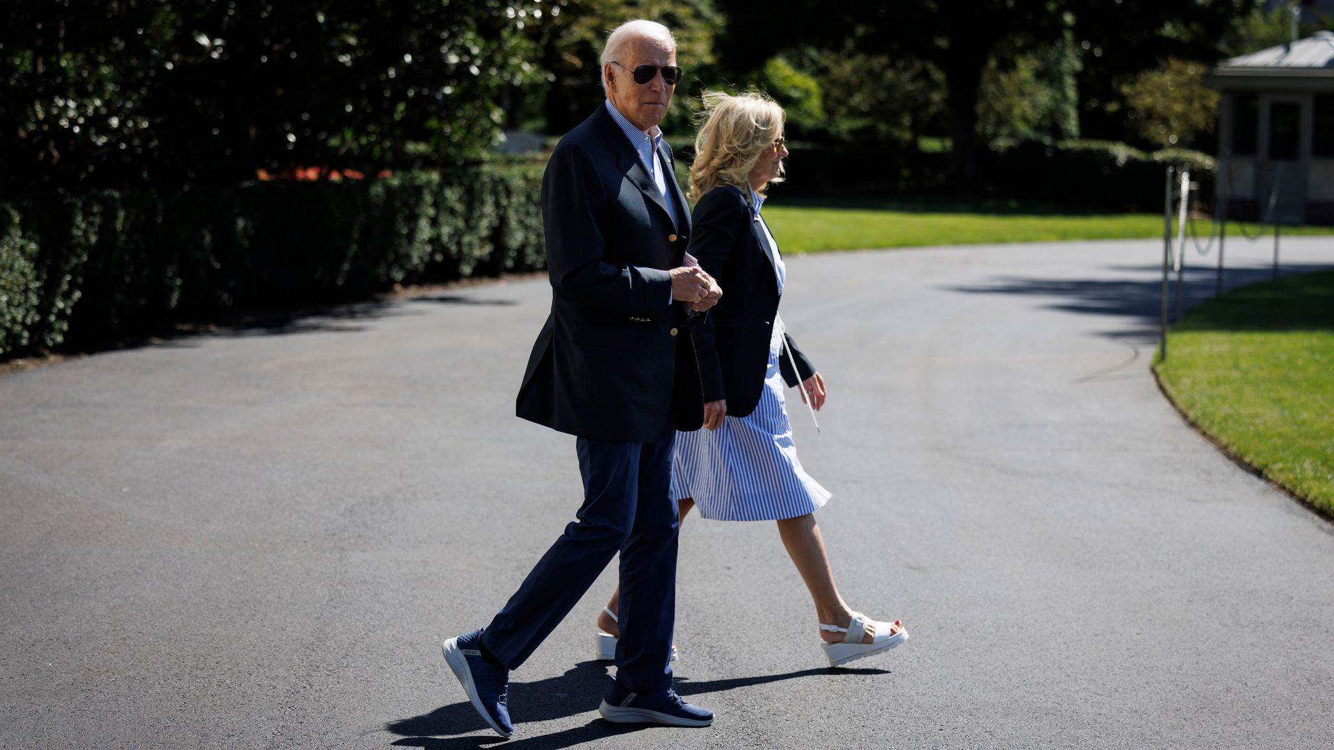 President Biden and First Lady Jill Biden walk across a driveway at the White House. The president is wearing a sports jacket, casual trousers and tennis shoes.