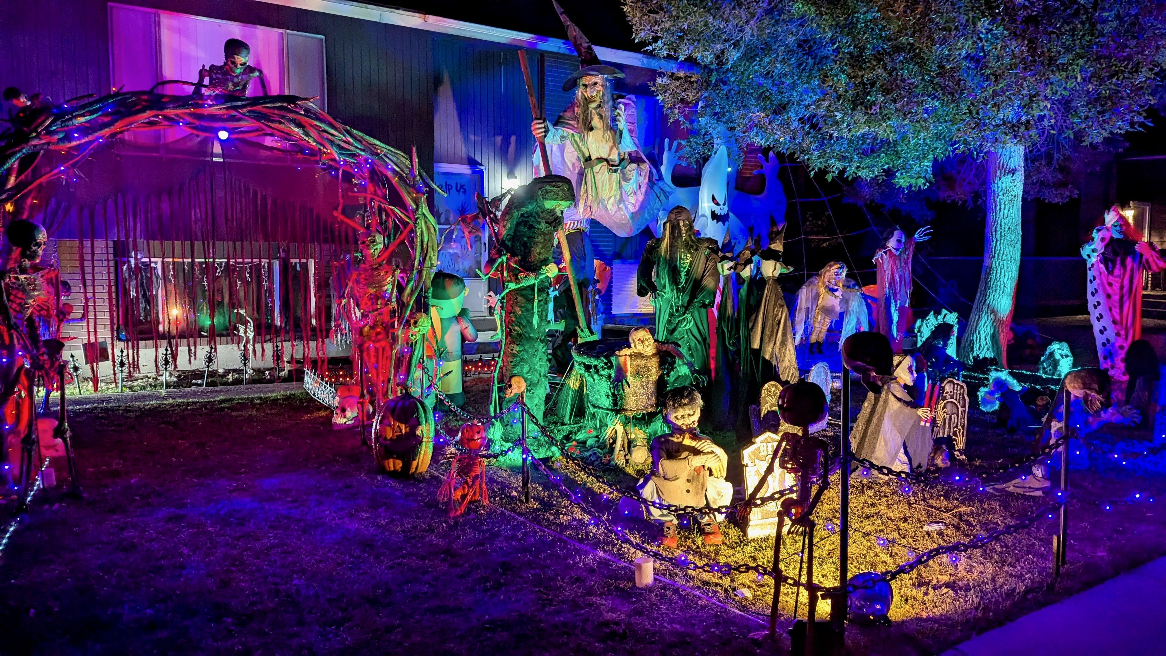 Nighttime Halloween front yard display with colorful neon lights, giant skeletons, witches, ghosts, pumpkins, and other spooky figures around a fence and trees.
