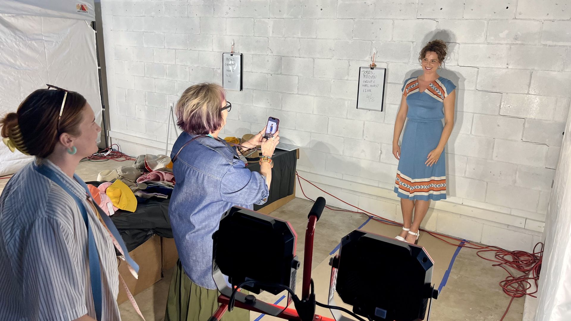 A woman in a blue dress with orange trim stands against a white brick wall, smiling as two people photograph her with a smartphone; studio gear, cables, and a cluttered set surround them.