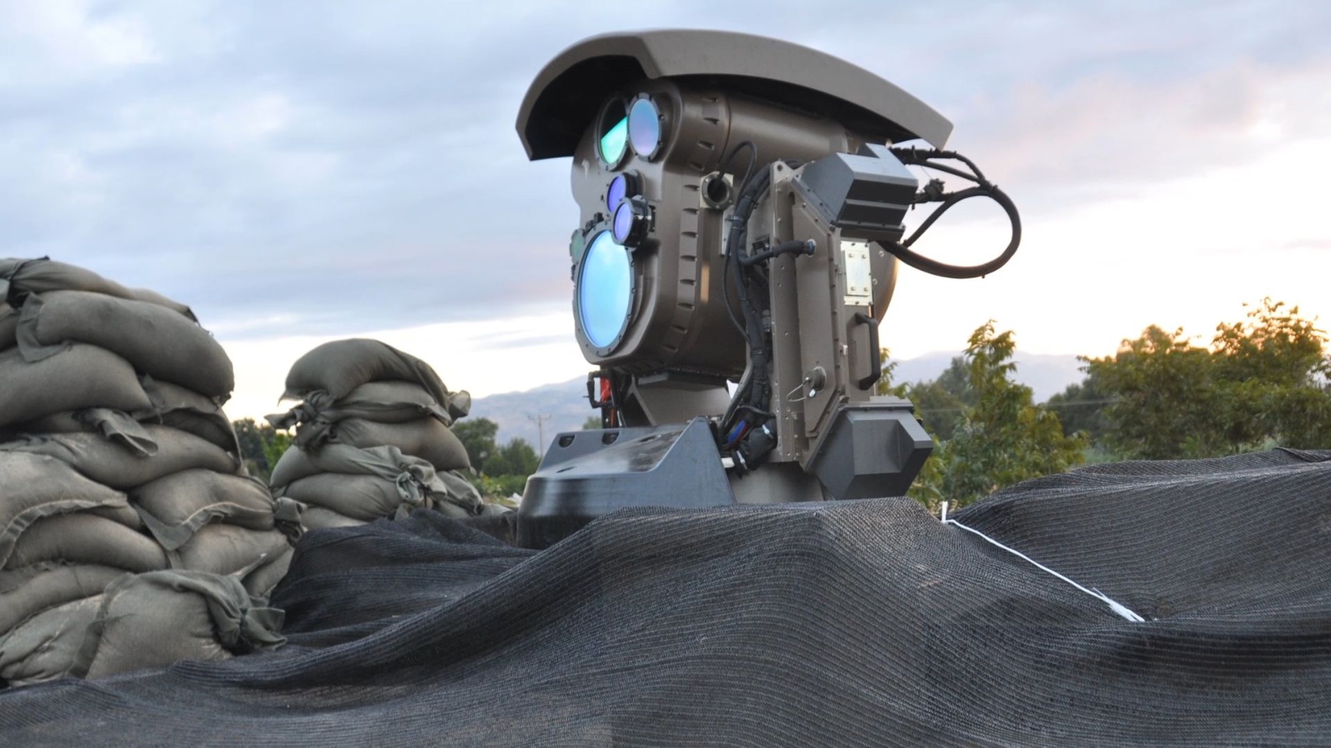 A laser weapon, mounted to a larger structure. Sand bags can be seen to the left of the frame. The sky looks like it' sunrise.