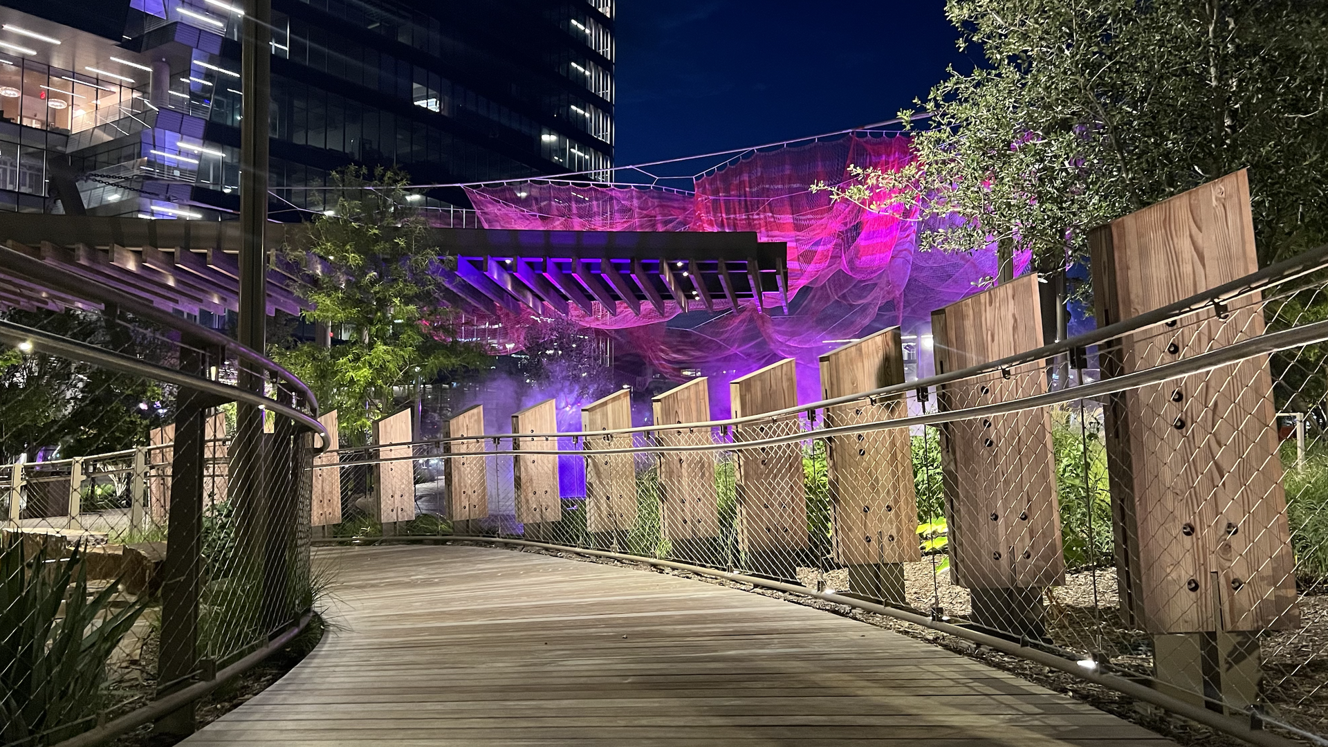 Night view of a curved wooden boardwalk with metal railing and vertical wooden panels, purple and pink illuminated netting overhead, trees, and modern building in background.