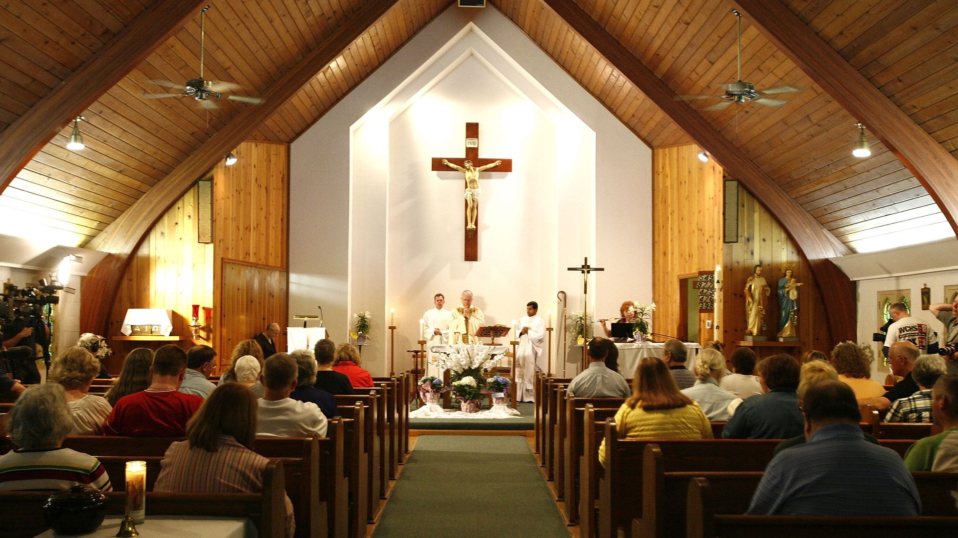 In this image, two rows of pews in a church are packed as church goers face a sermon being held in front of a white wall.