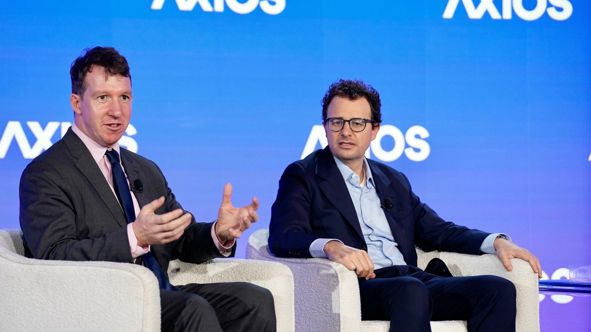 Two men in suits sitting in white armchairs on a stage with a blue background displaying the word AXIOS in white letters. One man gestures with hands, the other listens.