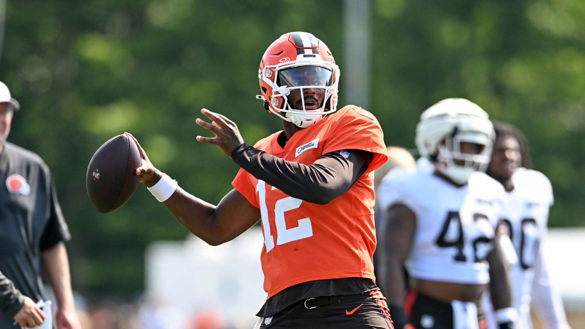 Football player in orange jersey #12 and red helmet throwing a football during practice, with teammates in white jerseys standing in the background on a sunny day.