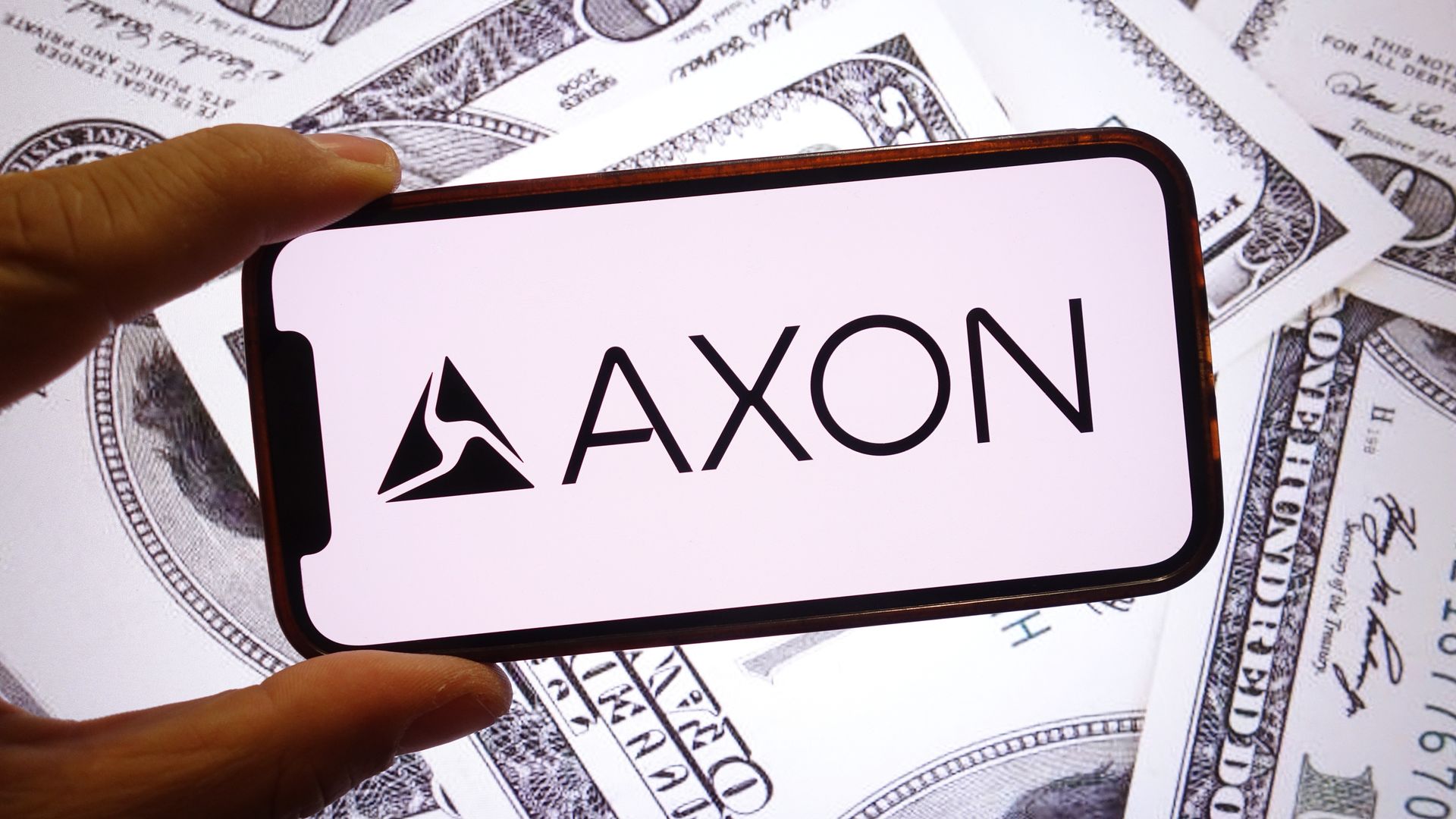 A hand holds a smartphone displaying the AXON logo with a triangle emblem, set against a backdrop of scattered US dollar bills.
