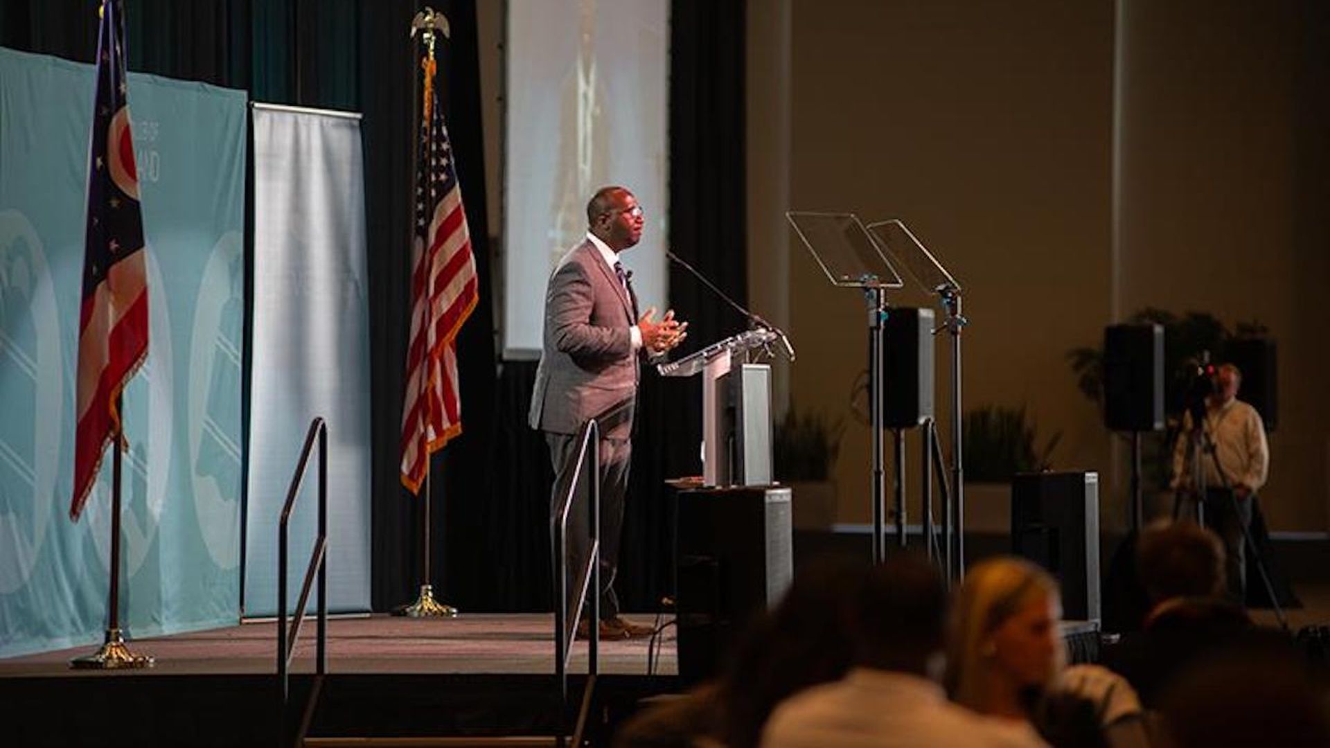 A bald Black gray-suited man in profile speaking at a lectern. 