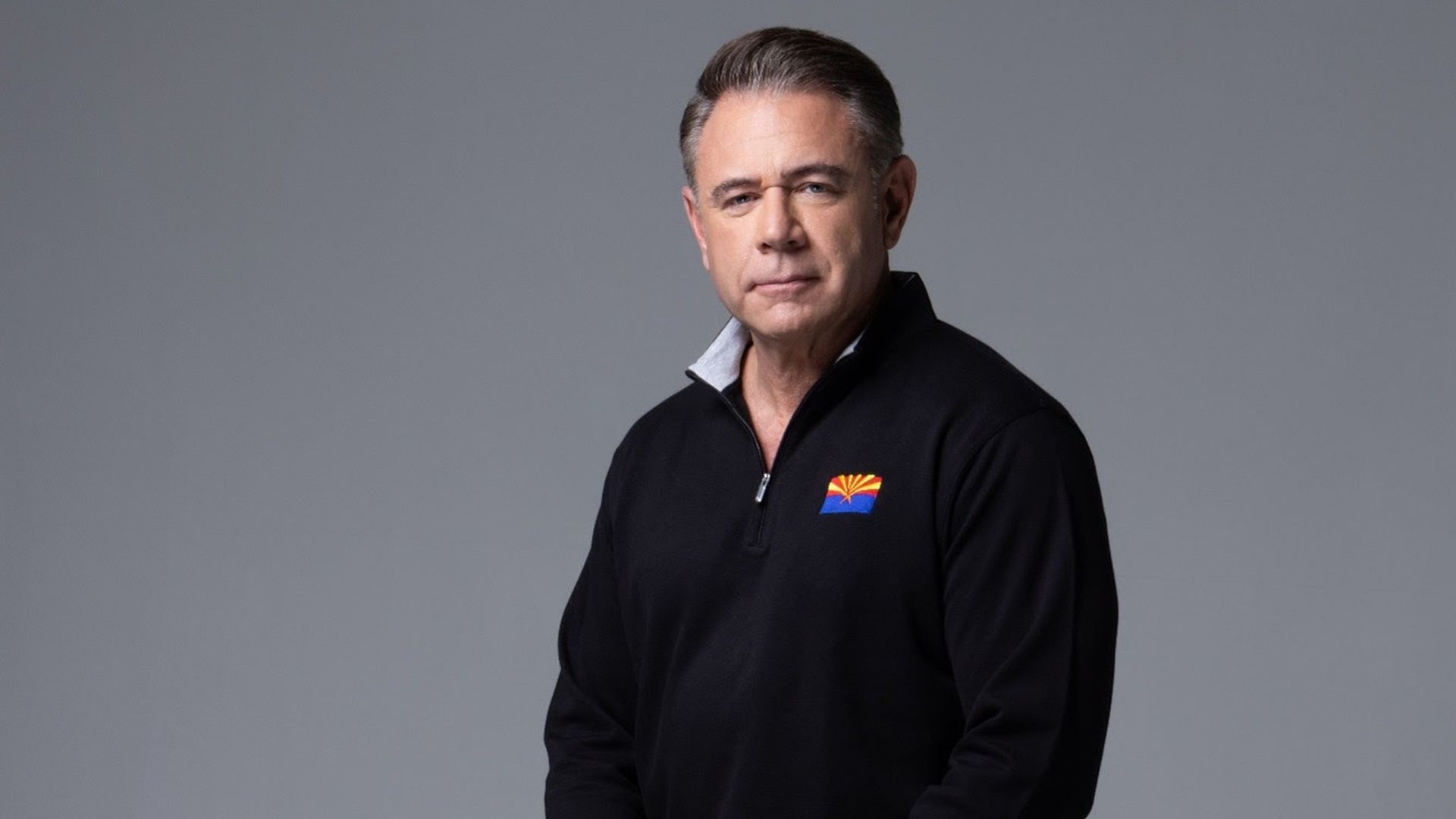 Middle-aged man with neatly combed hair, wearing a black quarter-zip pullover with a colorful Arizona state flag emblem, sitting against a plain gray background.