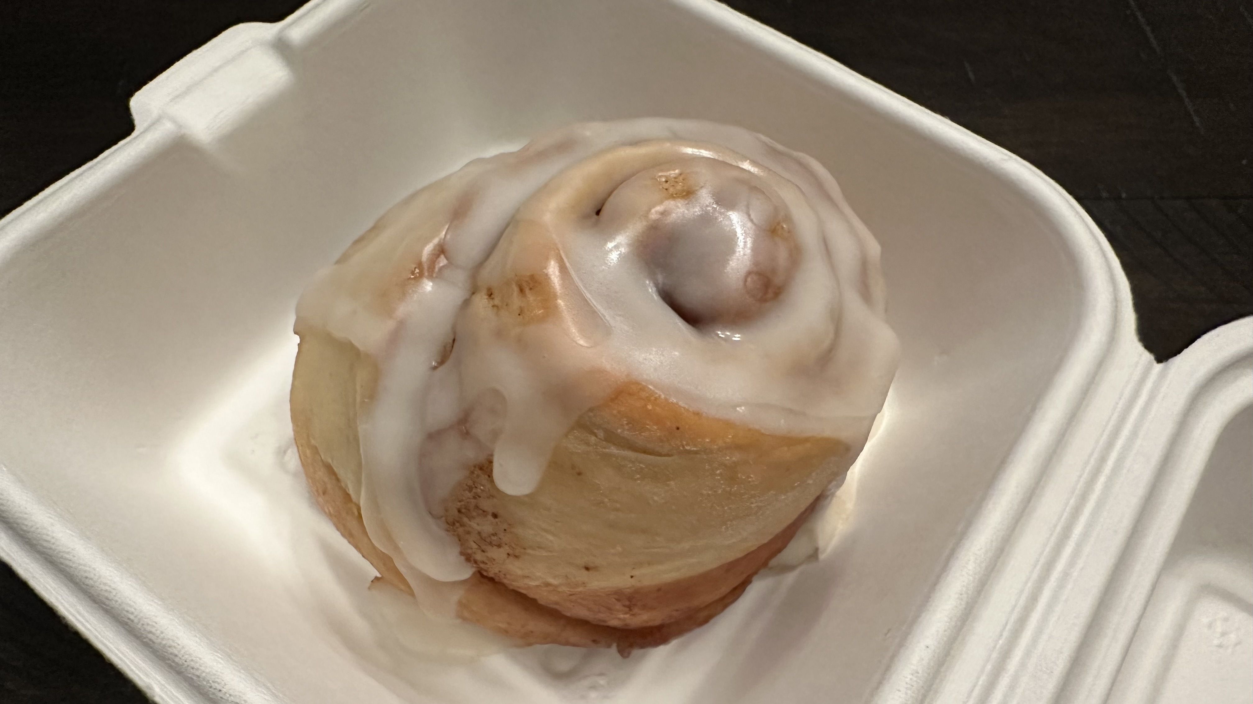 Close-up of a cinnamon roll with thick white icing in a white foam takeout container; the swirled pastry shows glaze and brown cinnamon filling.