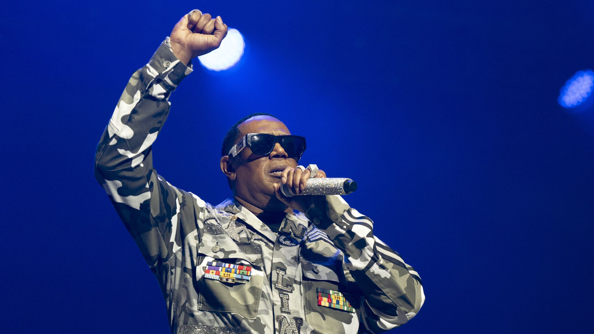 Male performer in grey and white camouflage jacket with medals, large black sunglasses, and glittery microphone, raising fist on stage with blue light background