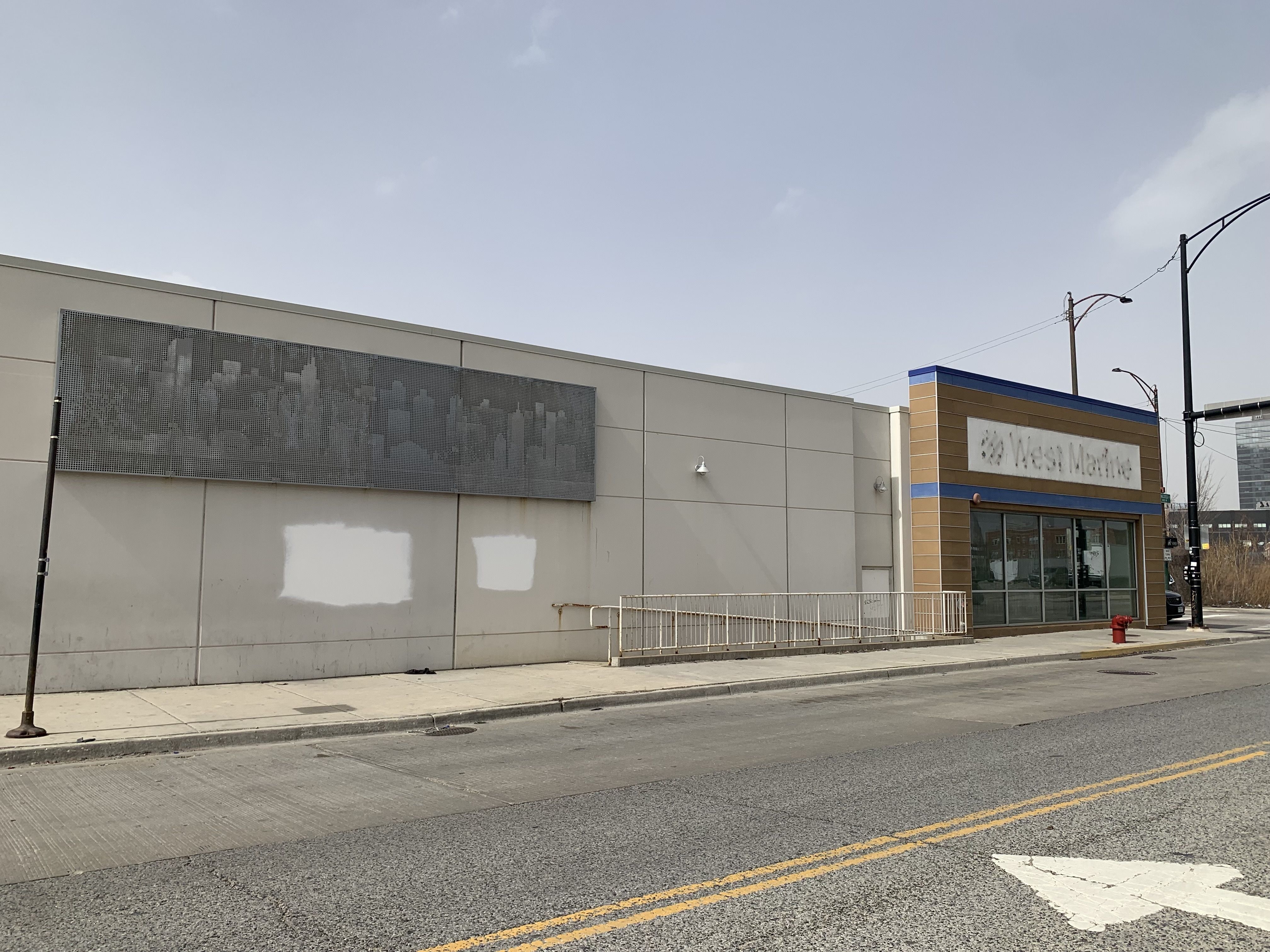 Empty street view of a beige building with a faded sign reading "West Market" and a large mural of a city skyline on the wall under a cloudy sky.