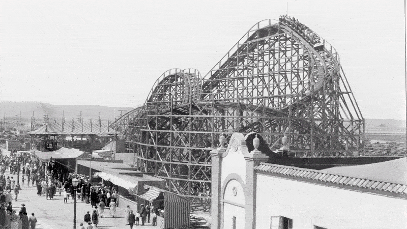 A historical photo of the Giant Dipper on Belmont Park's opening day in 1925. It dissolves into a colorful aerial photo of the roller coaster and beachside amusement park in present day at sunset. 