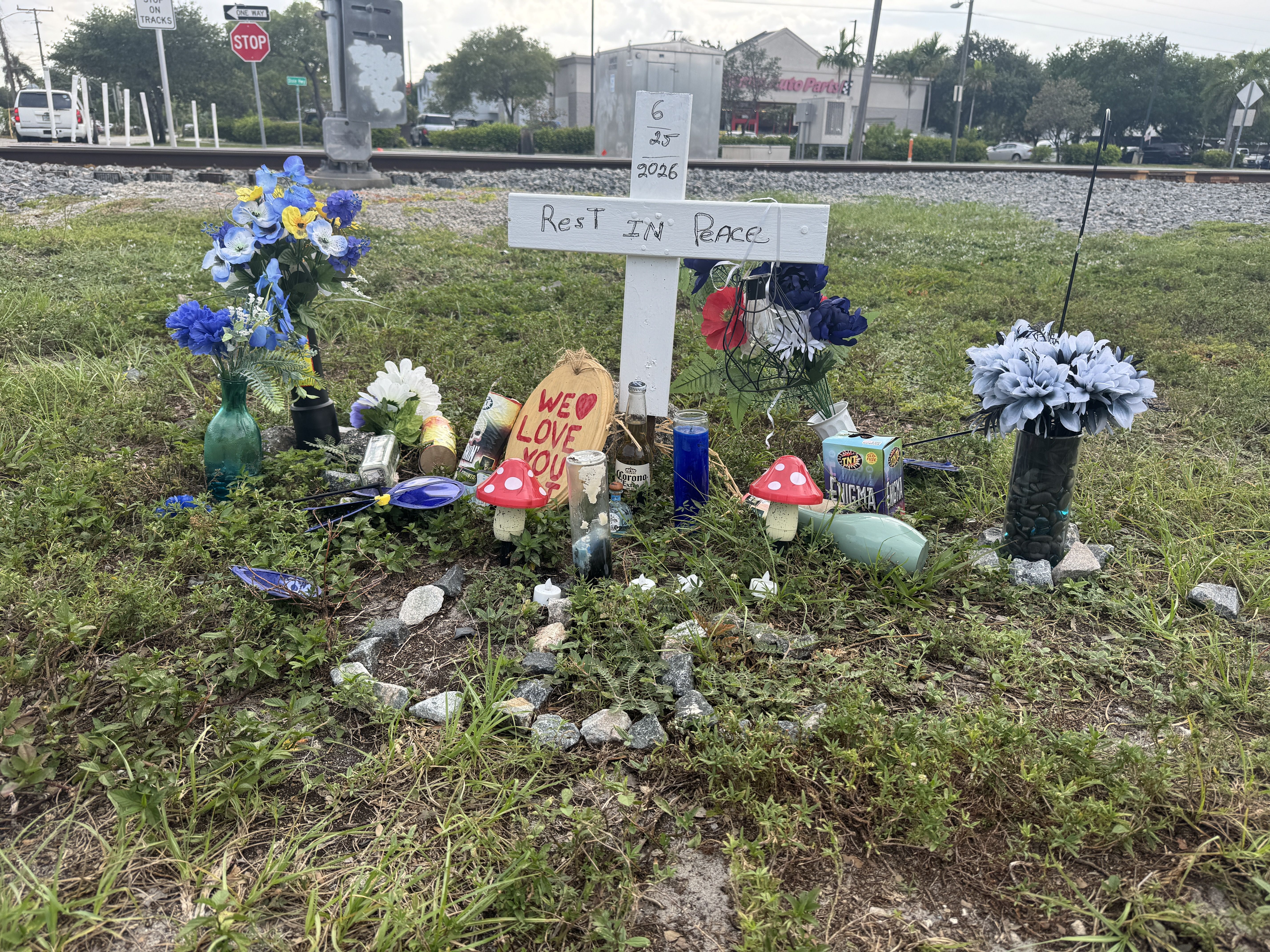 Roadside memorial by railroad tracks: a white cross with Rest In Peace and date 6-25-2026, surrounded by blue and yellow flowers, candles, and keepsakes.