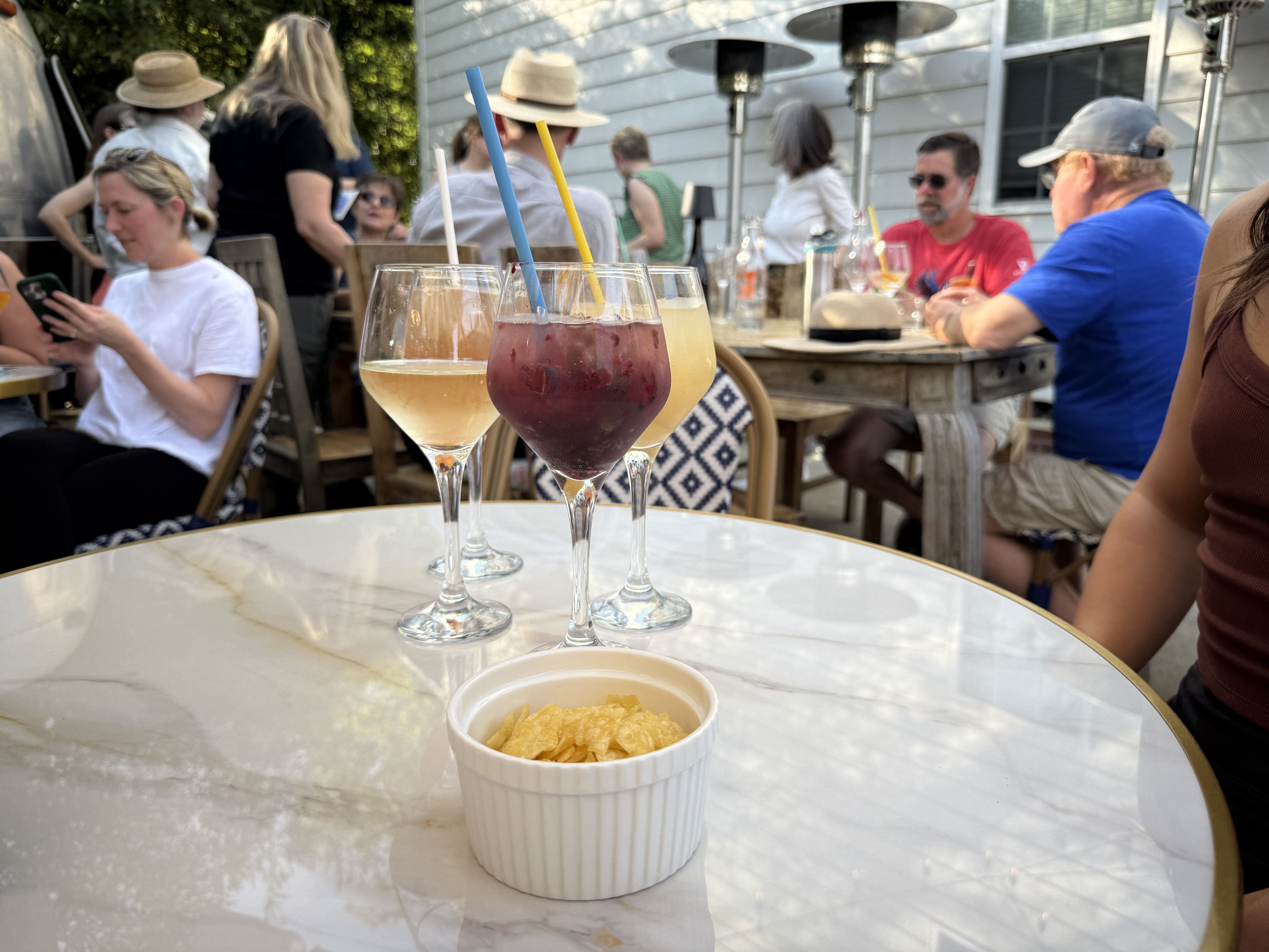 Outdoor cafe table with three stem glasses of pale yellow, pink, and purple drinks with blue and yellow straws, and a white ramekin of chips. In the background, people dine at wooden tables.