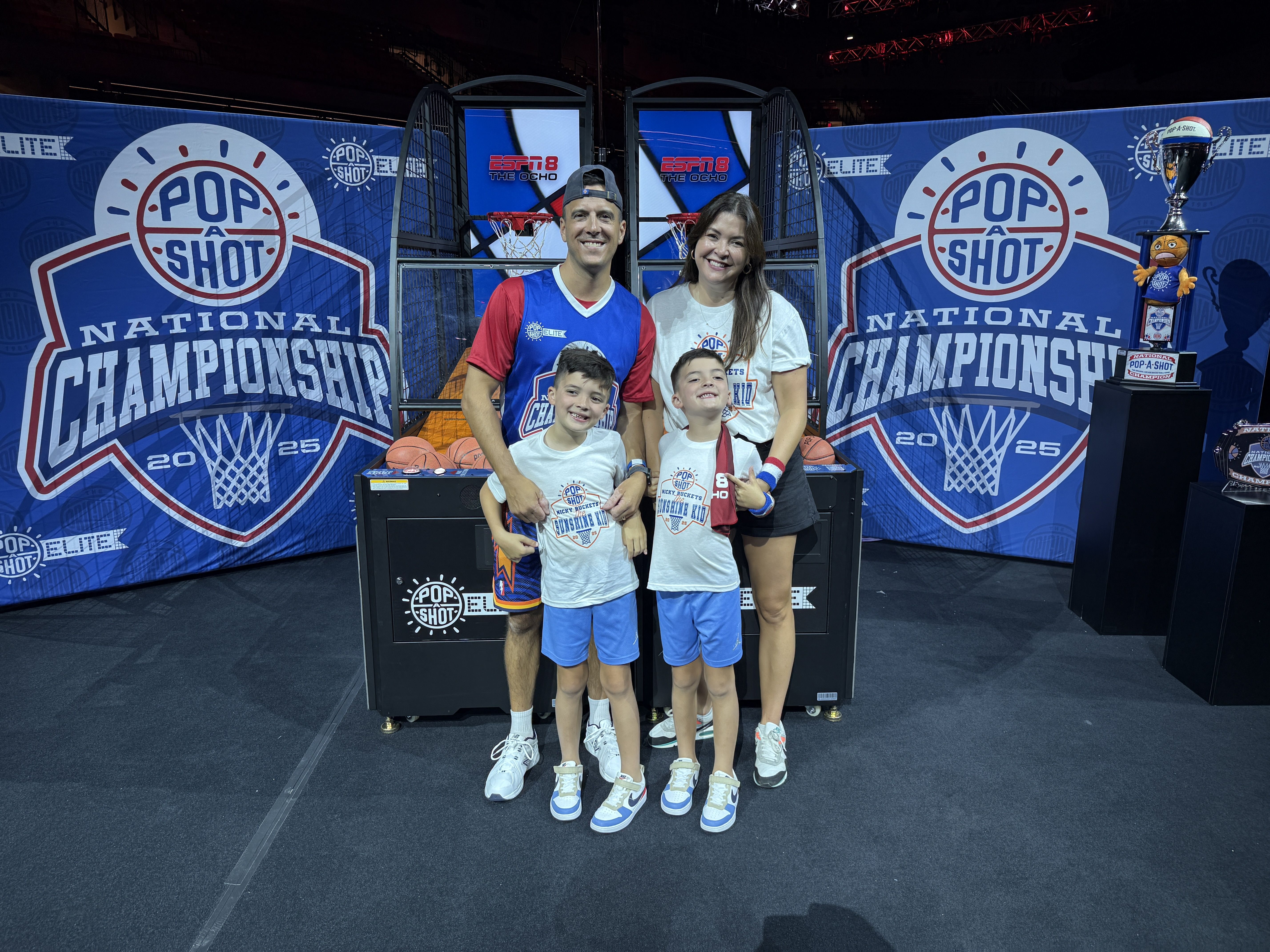 A smiling family of four poses in front of a "Pop-A-Shot" setup for the National Championship, with blue banners, two basketball hoops, and a trophy on the right.