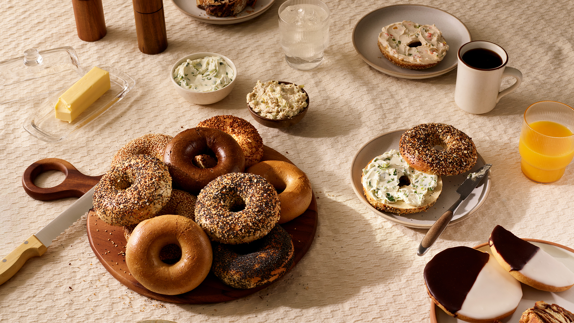 Table with assorted bagels on a wooden board, cream cheese spreads in small bowls, a butter stick with knife, two black-and-white cookies, coffee in a mug, orange juice, and glasses of water on a beige tablecloth.